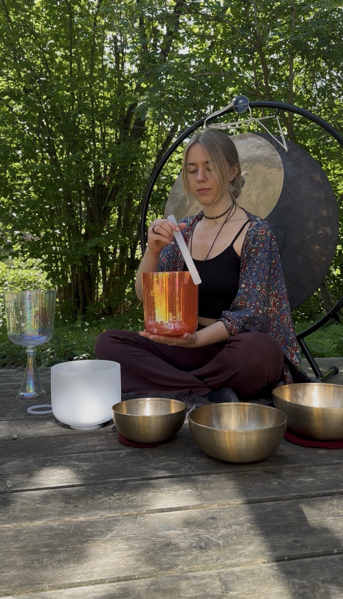 Frau sitzt im Schneidersitz im Freien und spielt Klangschalen und Gong zur Meditation.