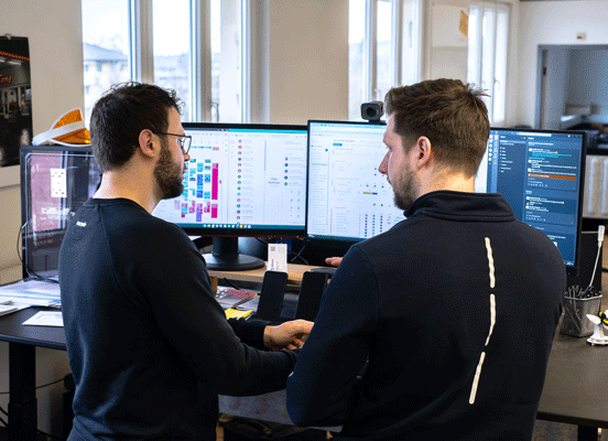 Zwei Männer besprechen sich vor drei Computermonitoren an einem Büroschreibtisch.