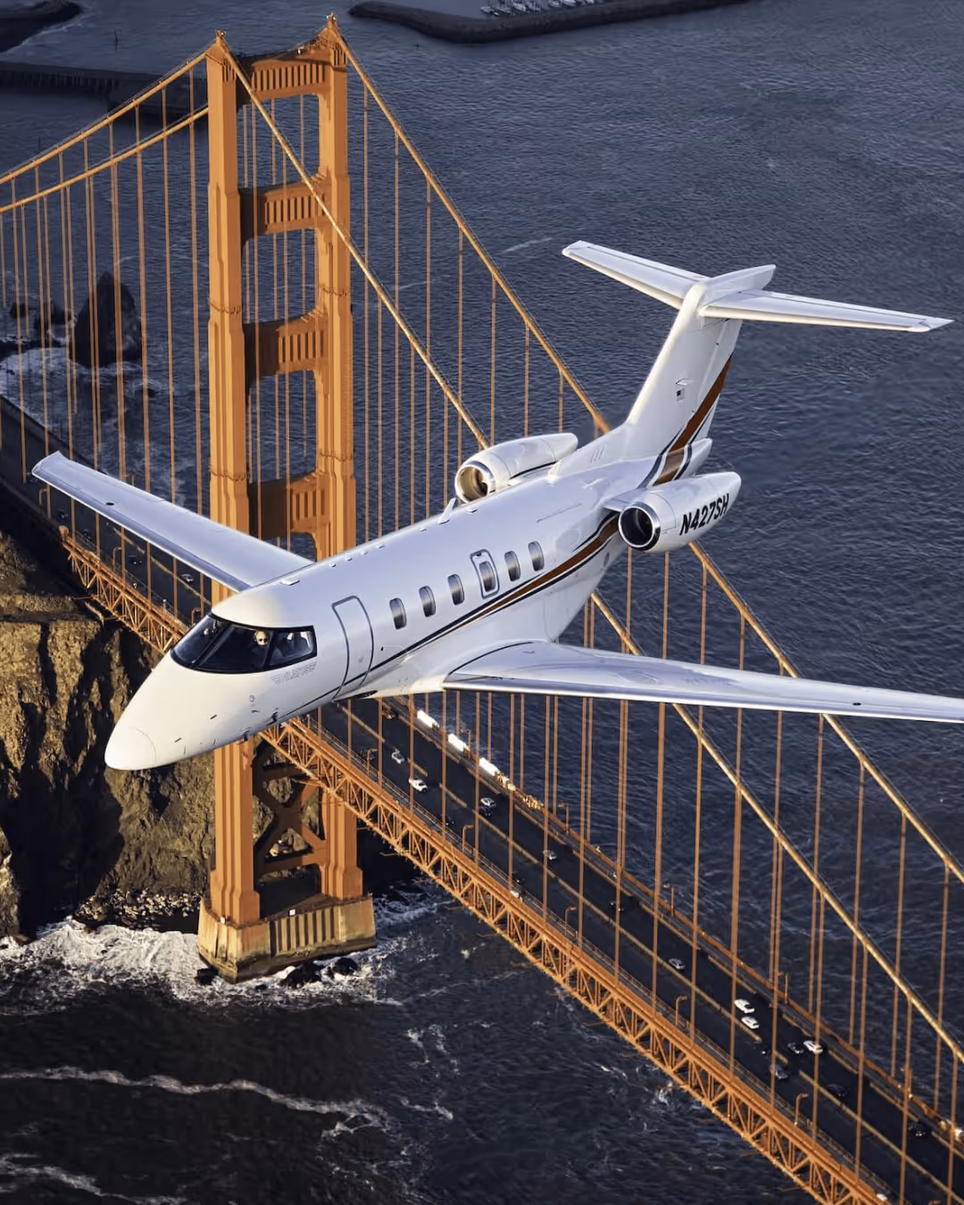 White private jet flying over the Golden Gate Bridge with water below.