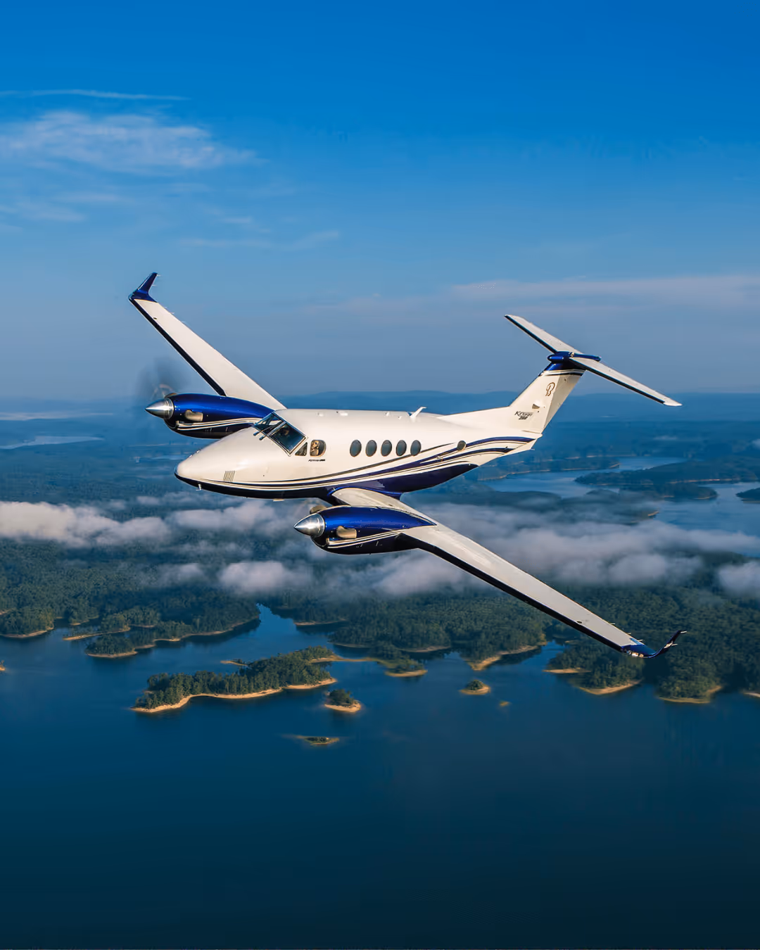 White and blue twin-engine propeller airplane flying over a lake with small forested islands under a clear blue sky.