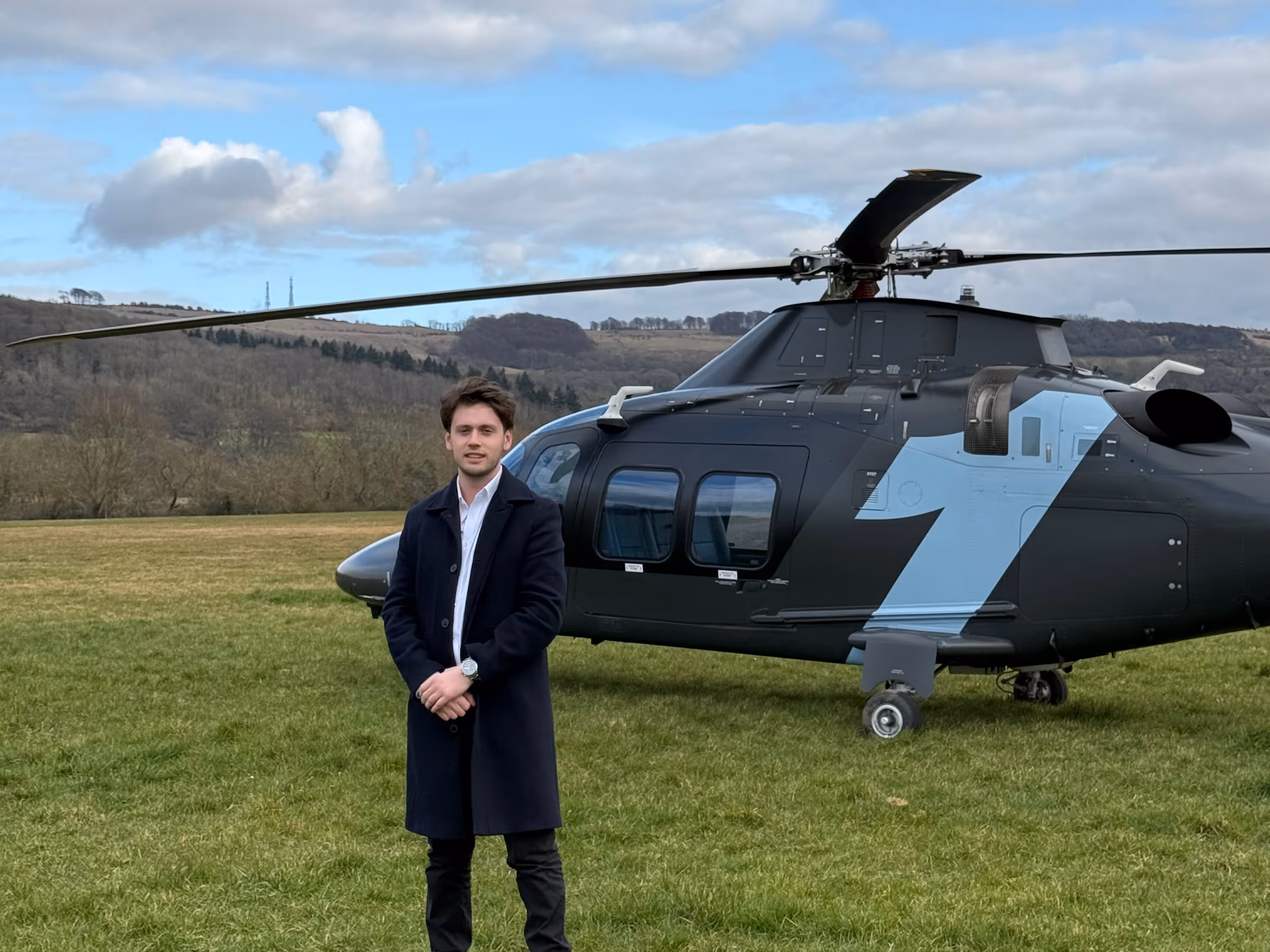 Young man in a dark coat standing on grass in front of a black helicopter with a light blue number one on its side, with a hilly landscape in the background.