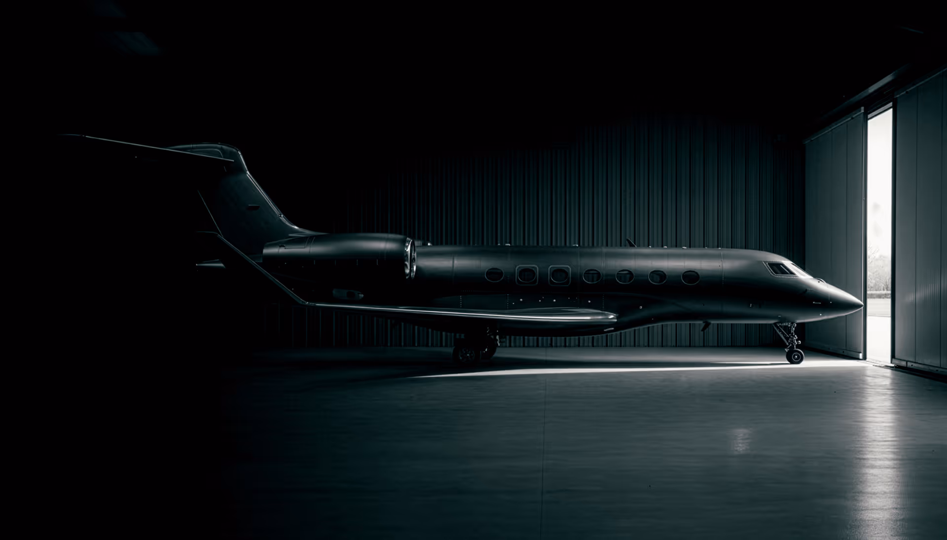 Private jet parked inside a dimly lit hangar with light coming from an open hangar door.