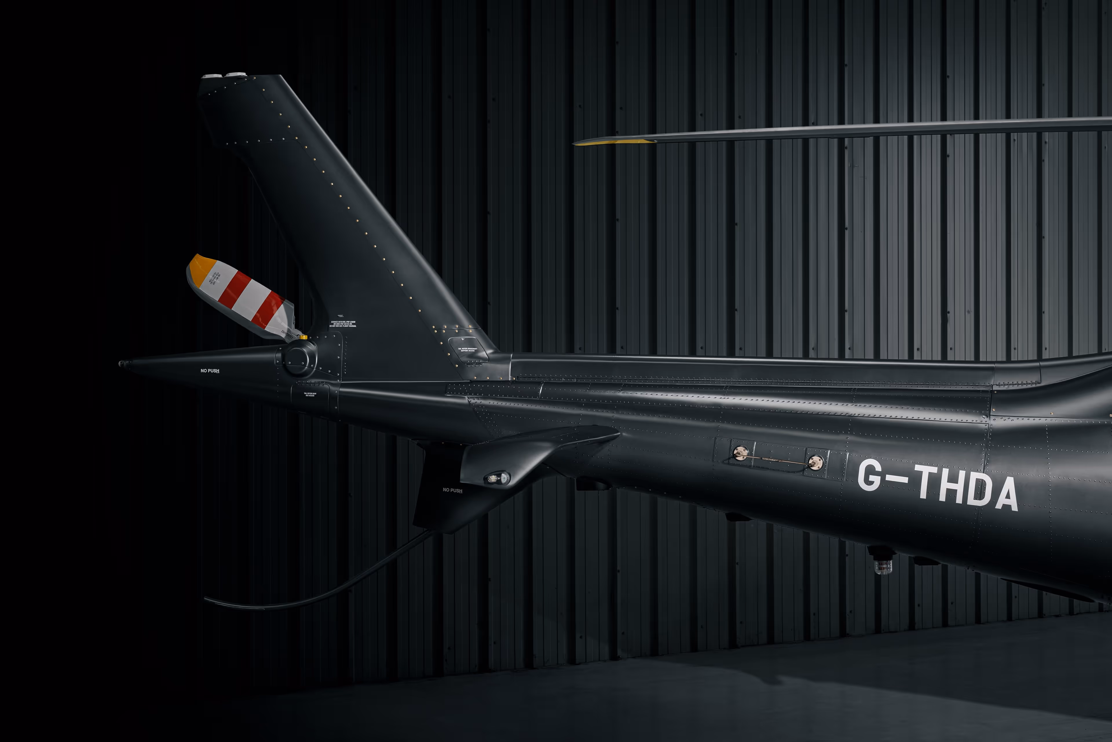 Close-up of a black helicopter tail with registration G-THDA in a hangar with corrugated metal walls.