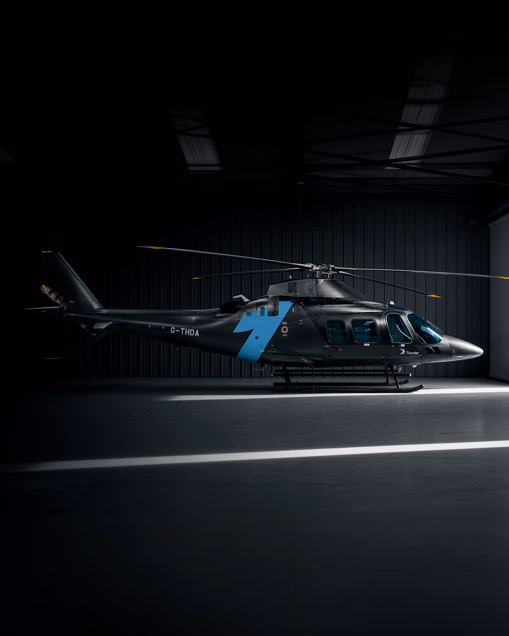 Black helicopter with blue lightning bolt inside a dimly lit hangar with light streaks on floor.