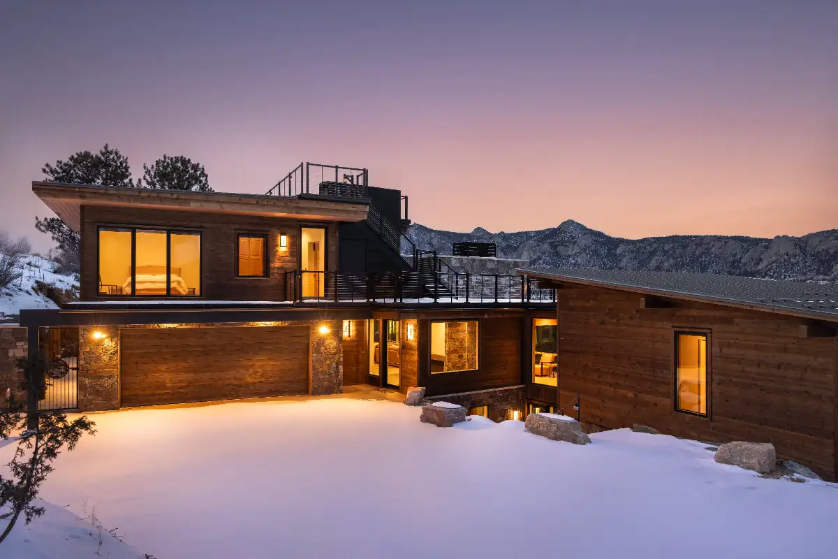 Mountain home exterior at dusk with Feeney cable railings on rooftop deck, wood and stone cladding, and snow-covered landscape