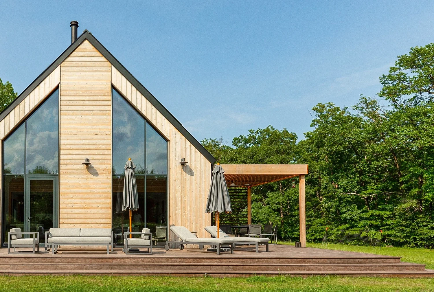 River Architects A-frame home with vertical wood cladding, full-height glass gable, outdoor lounge deck, and pergola set in woodland