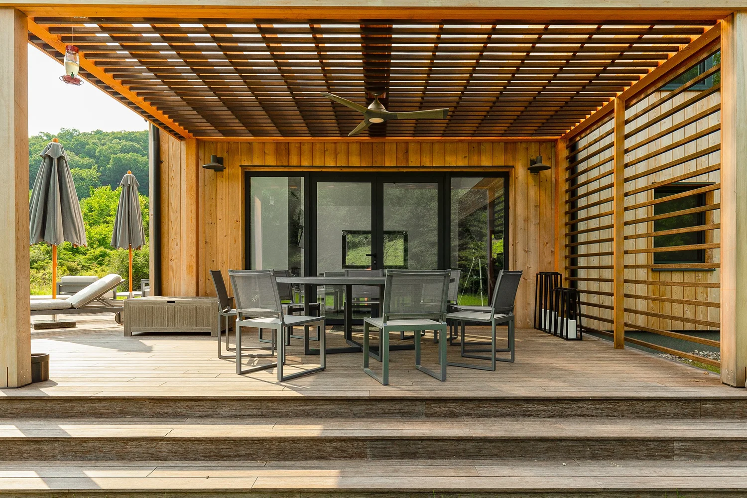 River Architects covered outdoor deck with wood slat pergola, dining set, and view into a modern timber-clad home