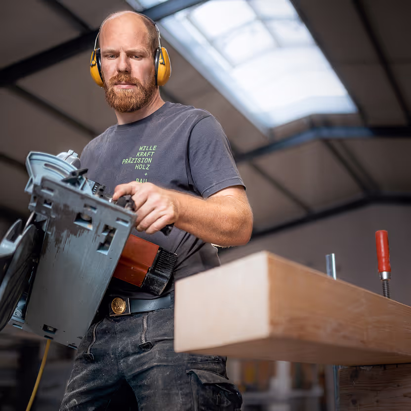 Ein Mann mit Ohrenschützern schneidet in einer Werkstatt mit einer Kreissäge einen Holzbalken.