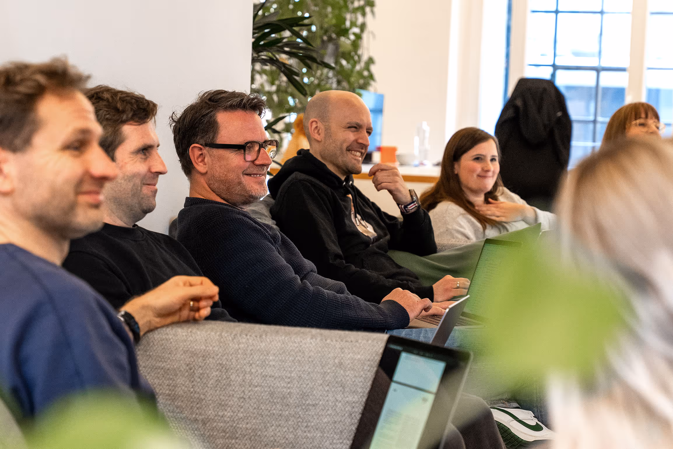 Eine Gruppe von Menschen sitzt auf Sofas mit Laptops und lächelt während eines Treffens in einem hellen Raum.