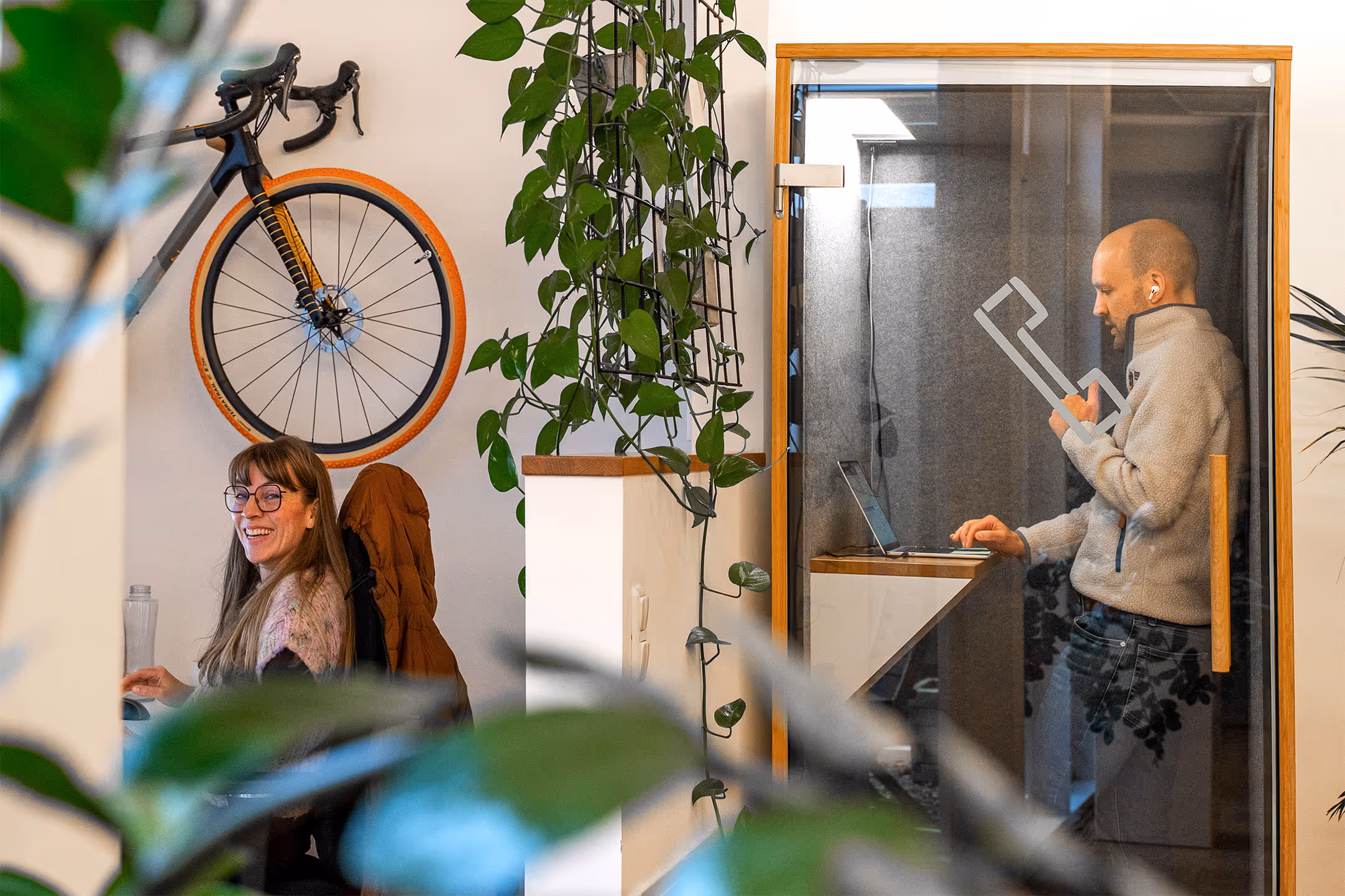 Frau mit Brille, die lächelnd an einem Schreibtisch sitzt, und Mann mit AirPods, der in einer kleinen Glasarbeitskabine an einem Laptop steht.