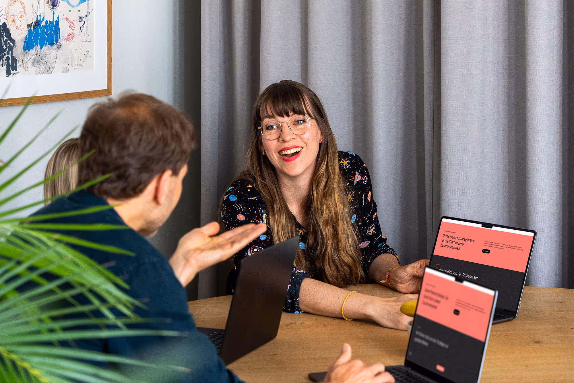 Zwei Personen im Gespräch an einem Holztisch mit zwei Laptops, eine Frau lächelt und trägt eine Brille.