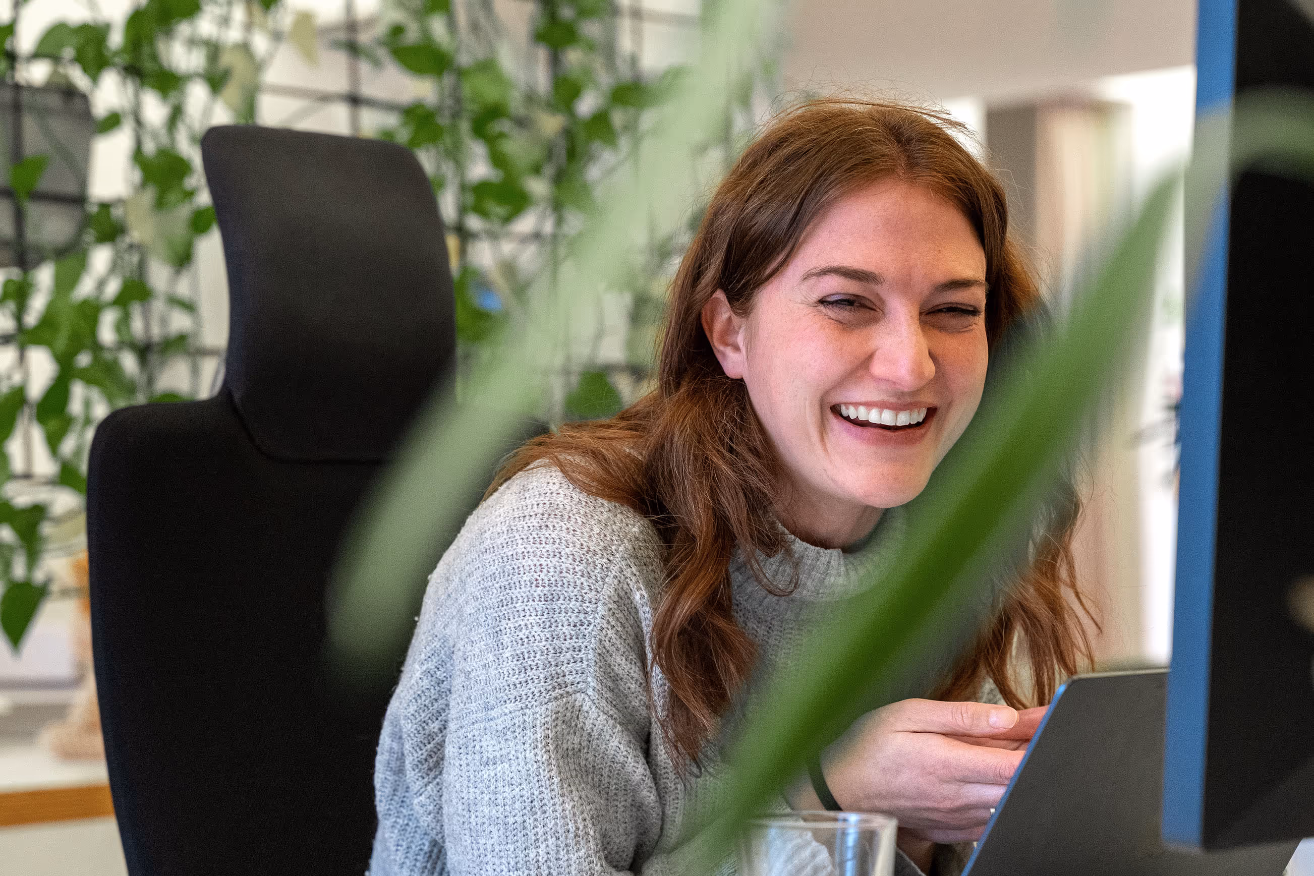 Lachende Frau sitzt an einem Schreibtisch vor einem Computer, umgeben von grünen Zimmerpflanzen.