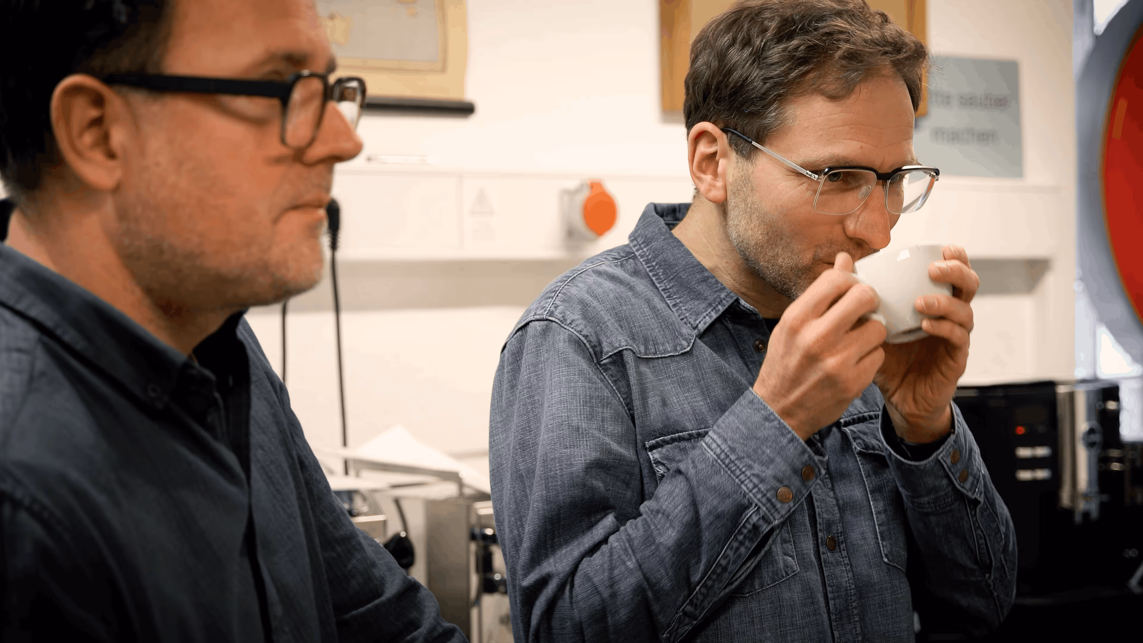 Zwei Männer mit Brille in Jeanshemden in einer Küche, einer trinkt aus einer weißen Tasse.