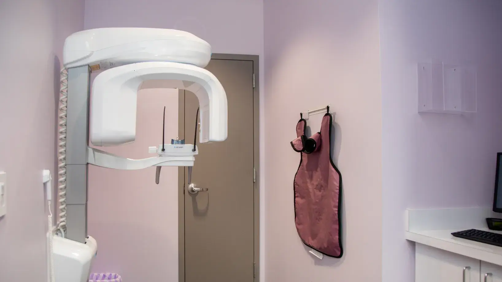 A dental X-ray machine in a dentist's office, next to a lead apron hung on the wall.