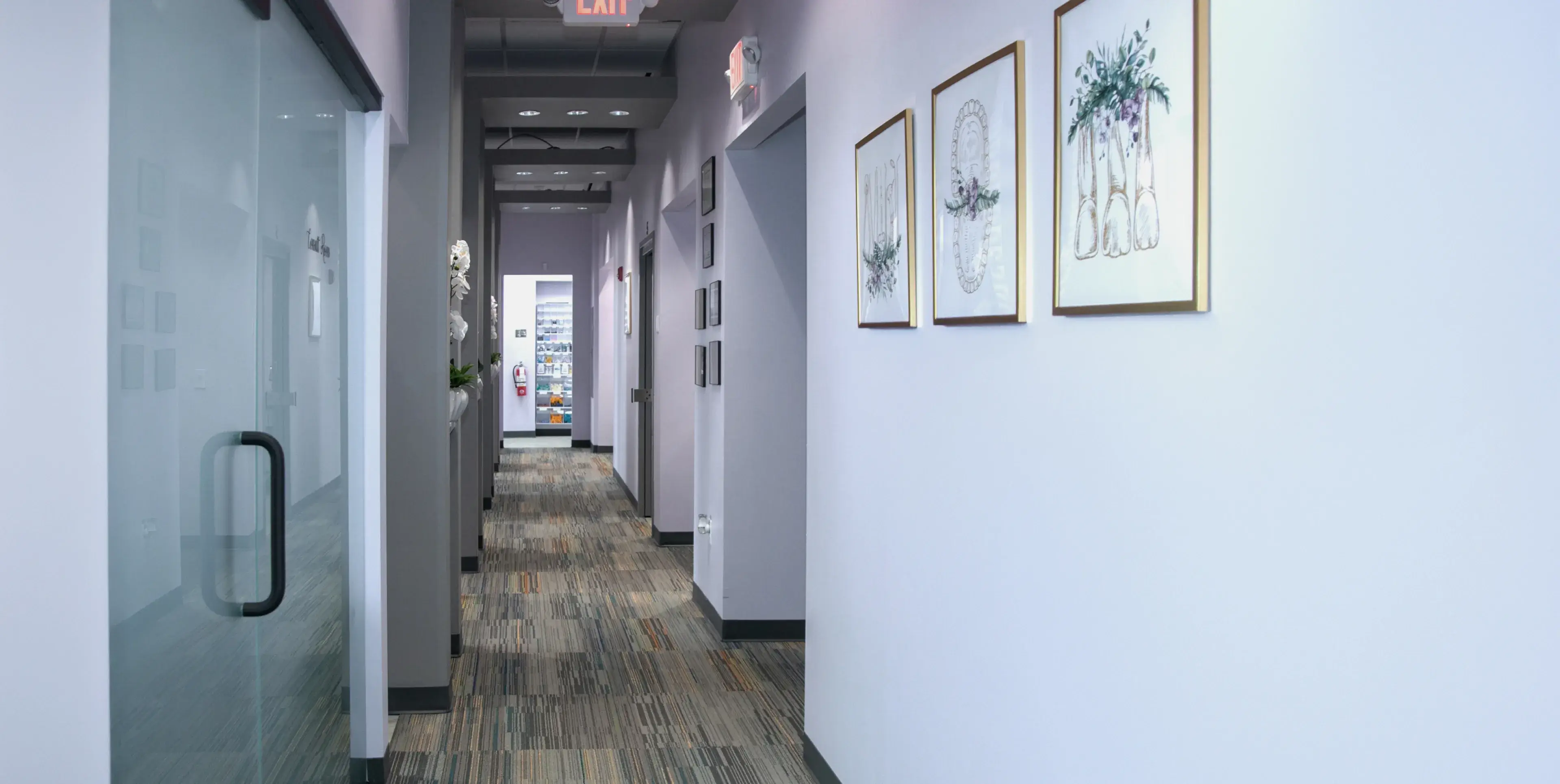A long hallway with framed pictures on the wall and an exit sign overhead.