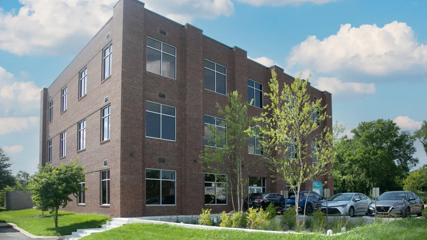 A three-story brick office building with cars parked in front on a sunny day.