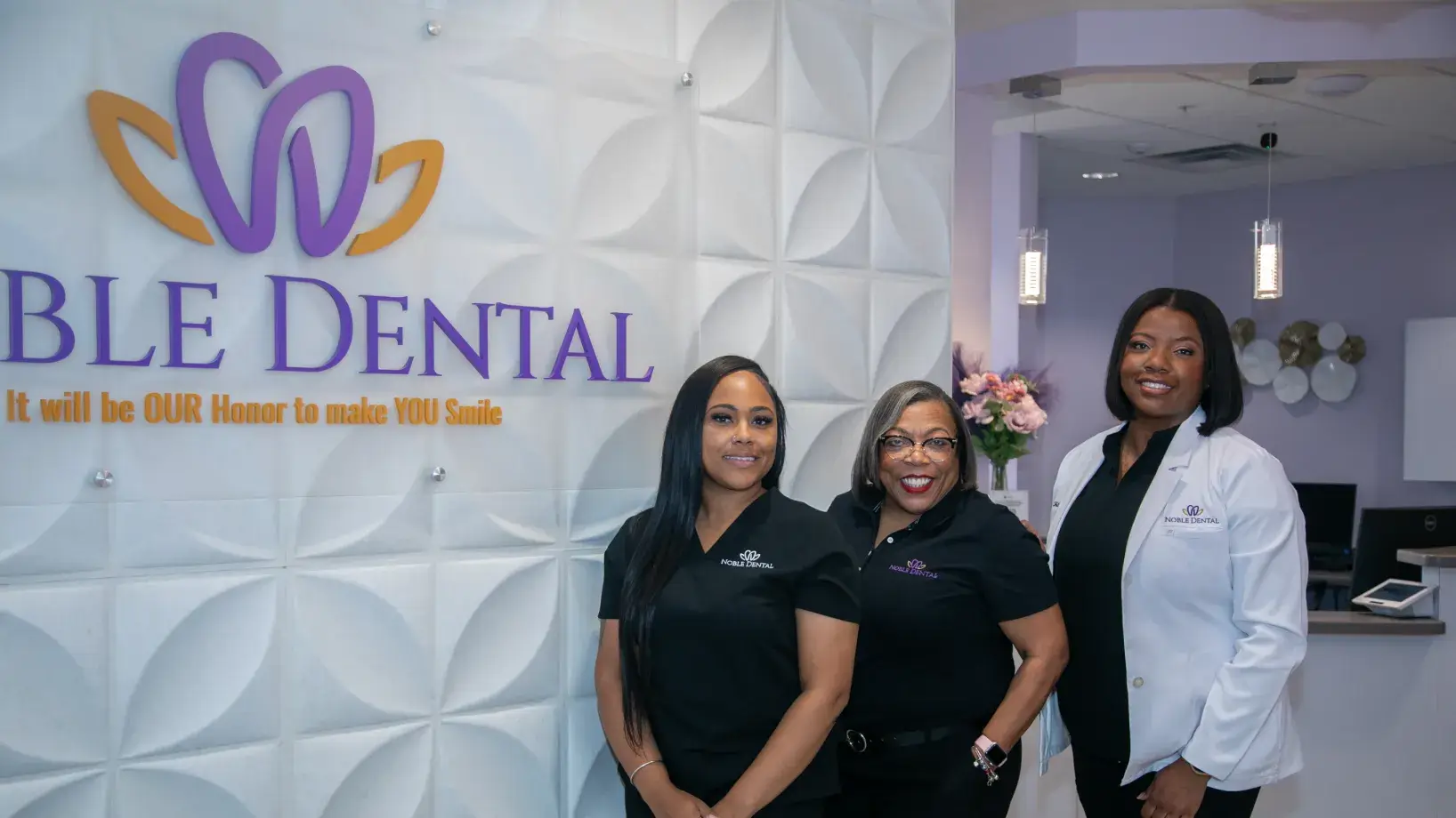 Three dental staff members in uniform smile in a dental office lobby near the reception desk.