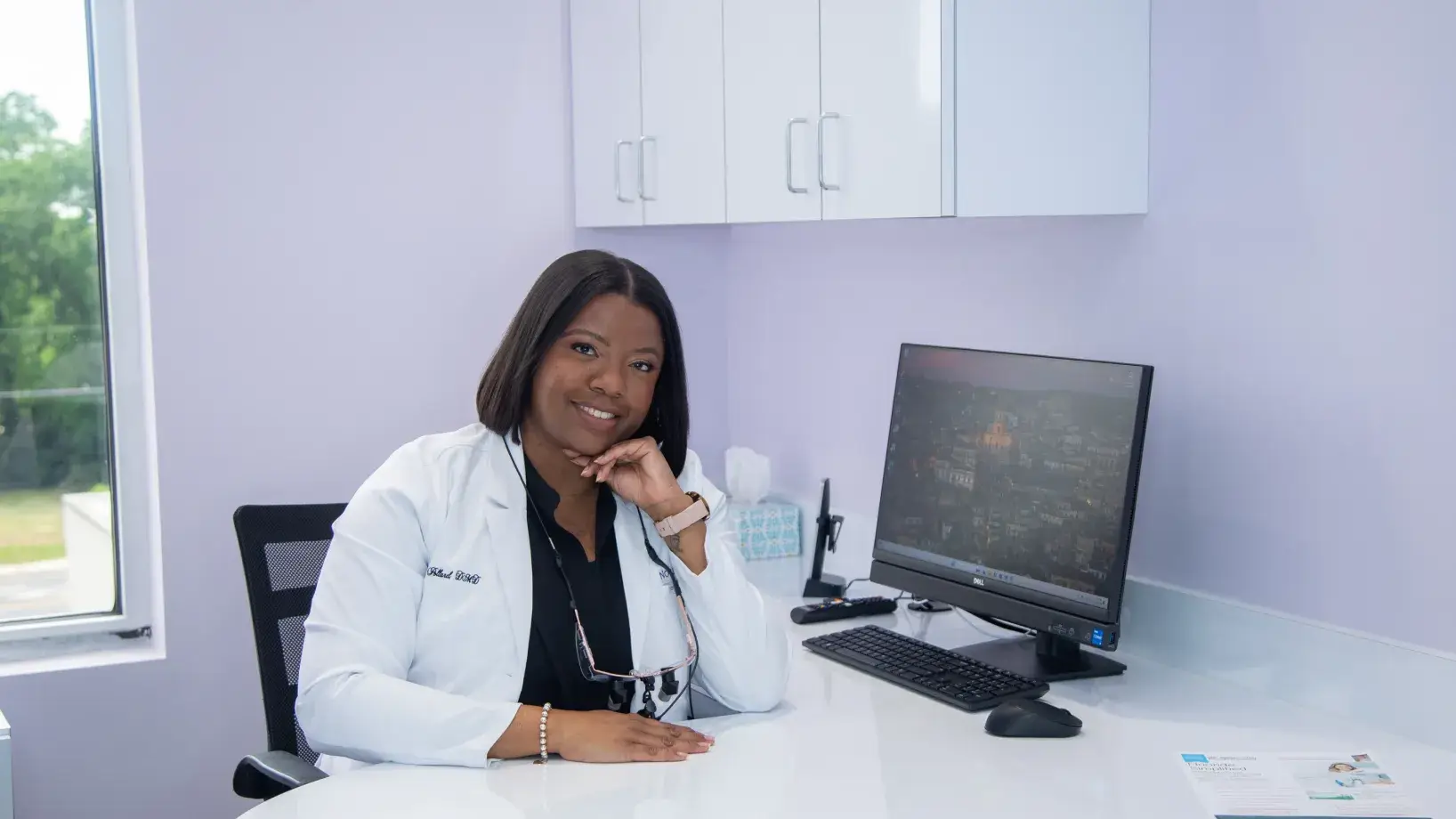 A woman in a white coat sits at a desk in an office, smiling at the camera.