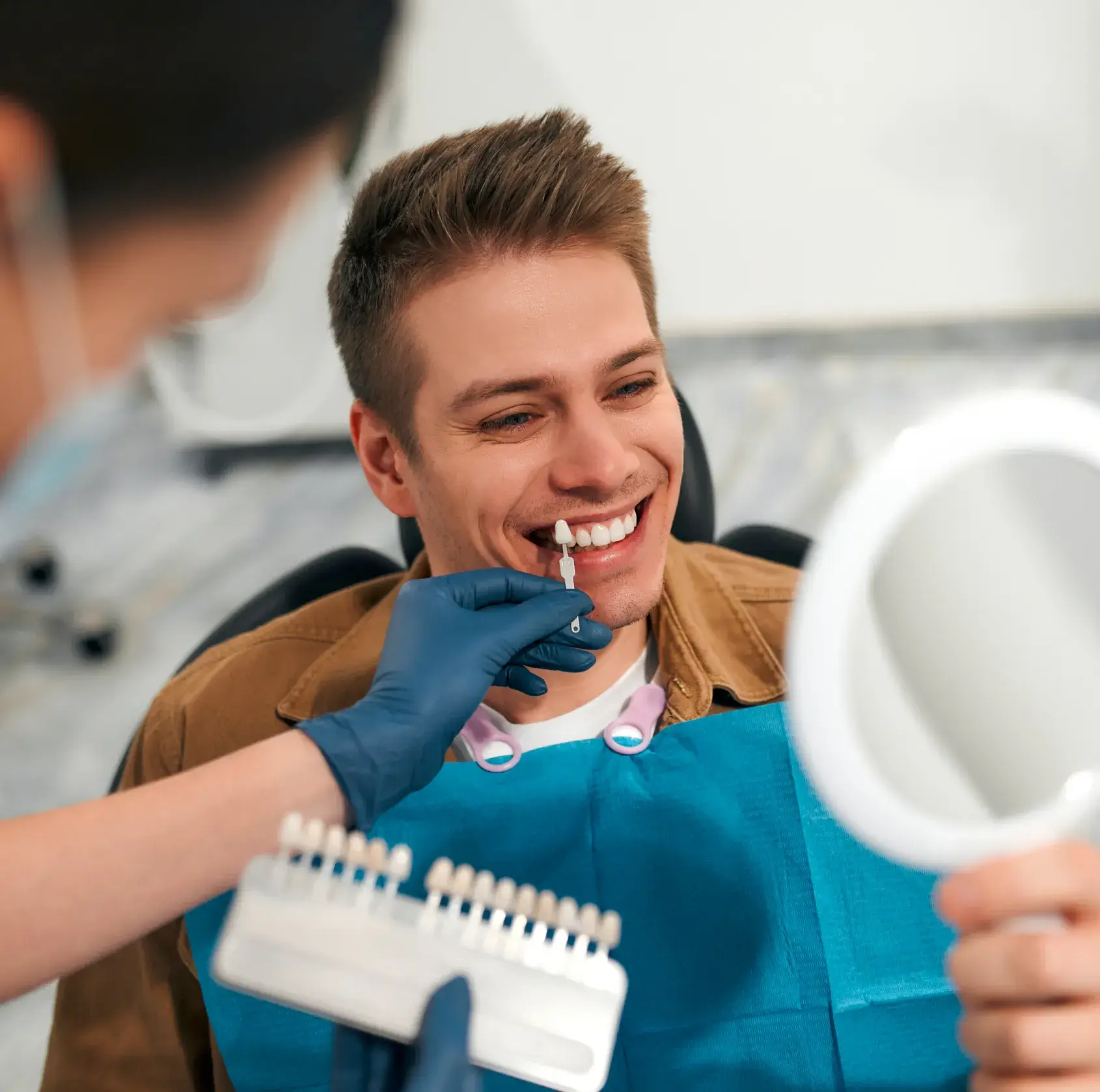 Dentist compares patient's teeth to a shade guide while the patient smiles, holding a mirror.