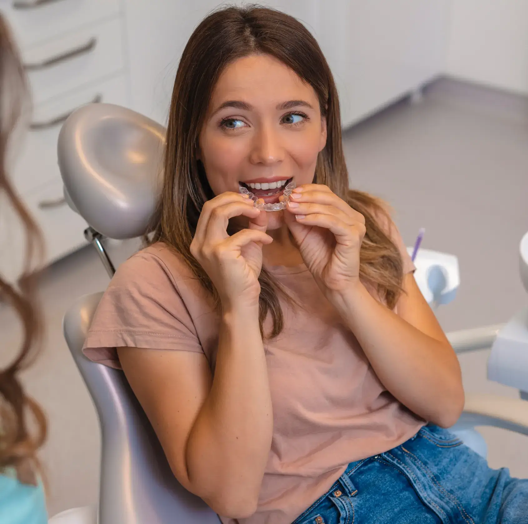 A woman in a dental chair is holding an orthodontic retainer near her mouth.