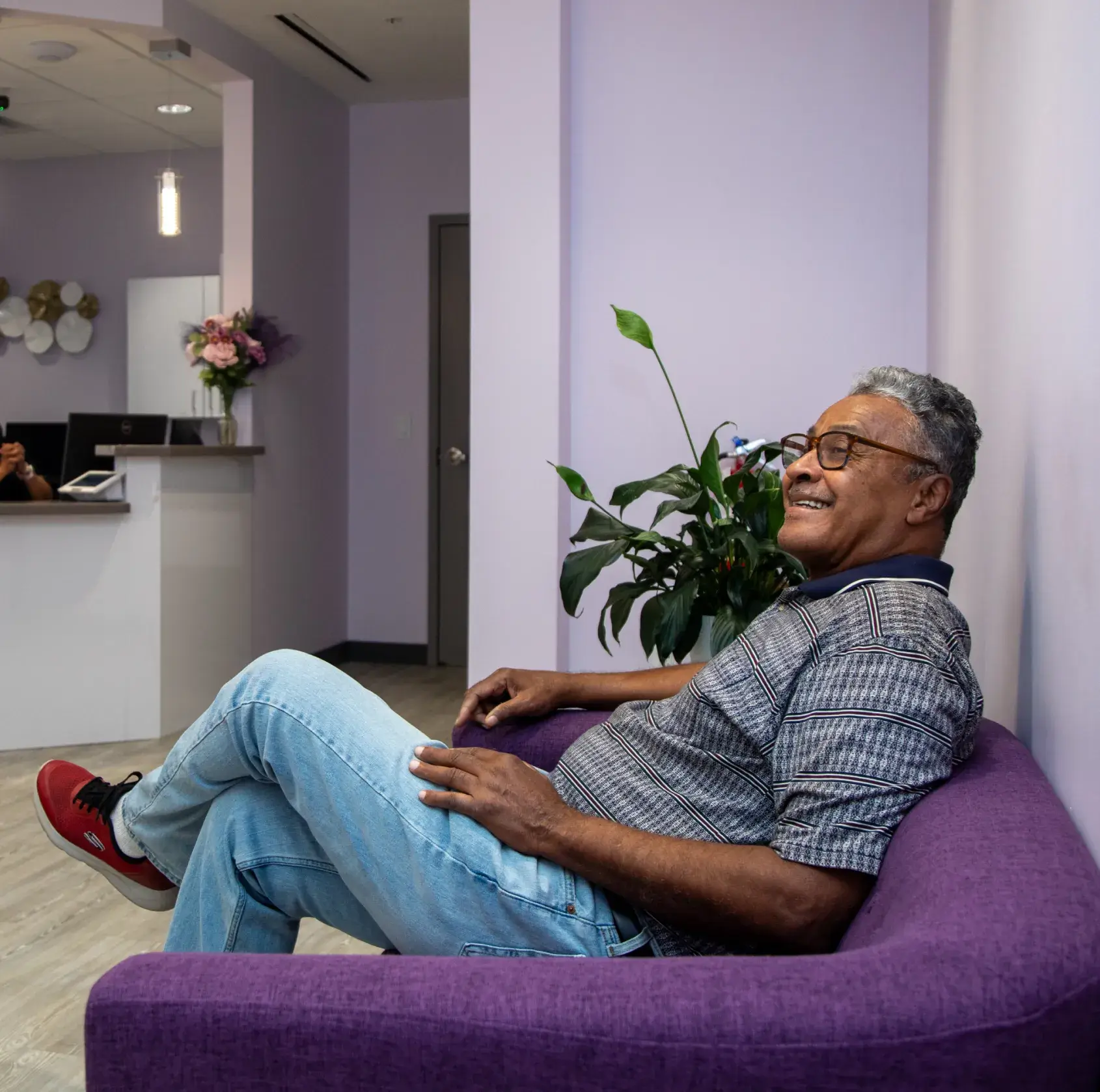 Man sitting on a purple couch, smiling, in a room with plants and a desk.