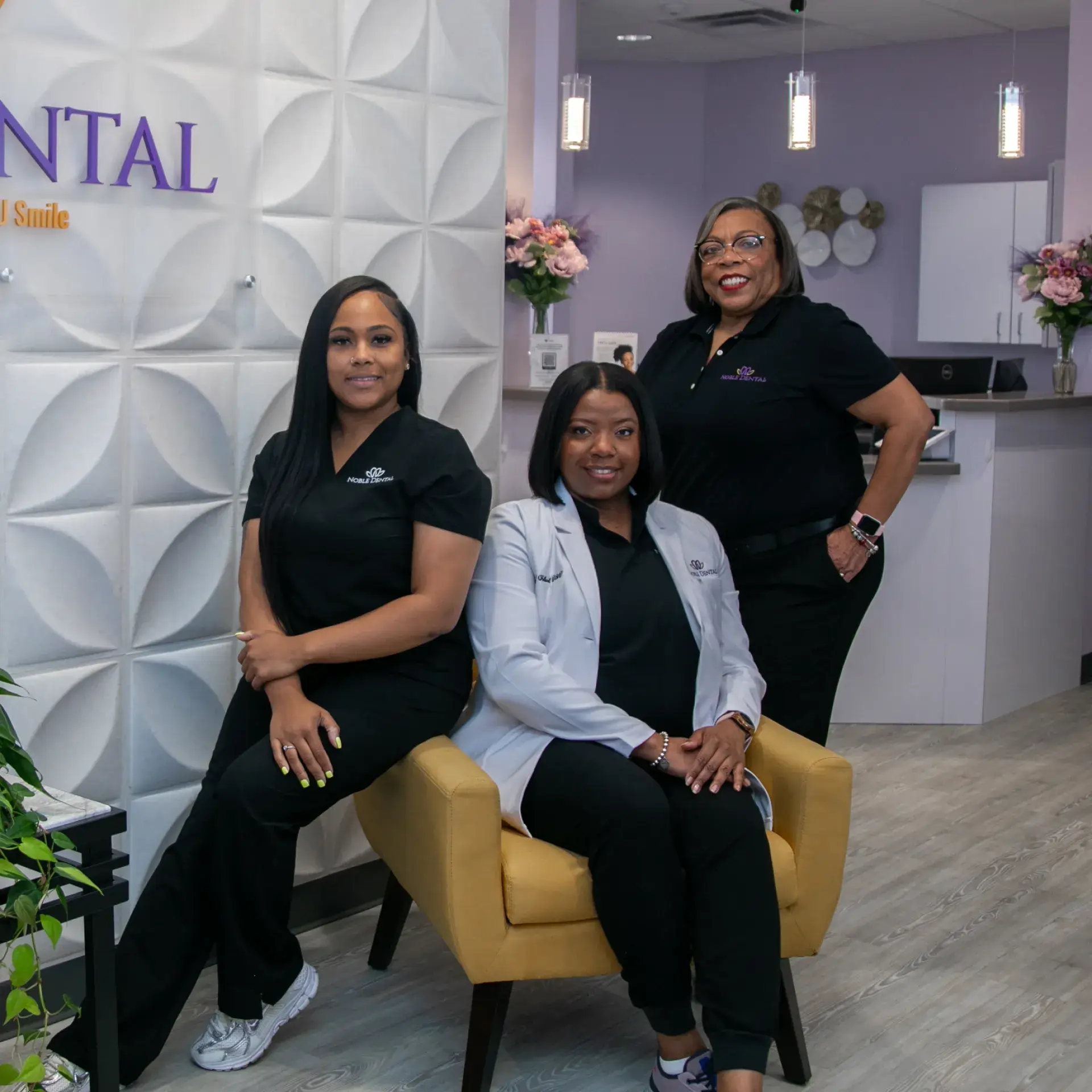 Three women in dental uniforms smiling, standing and sitting in a dental office reception area.