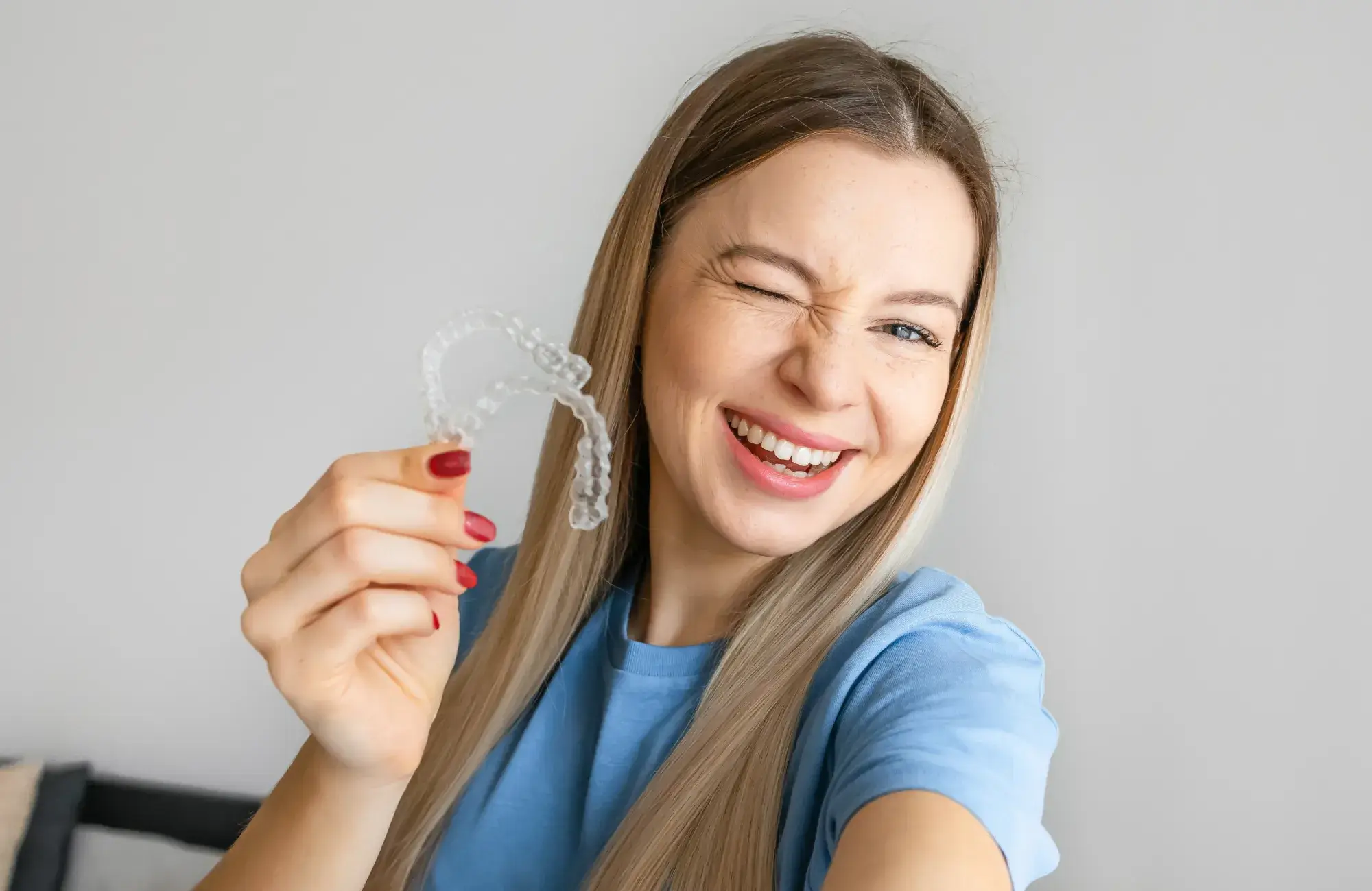 Woman smiling and winking while holding a clear dental retainer in her hand.
