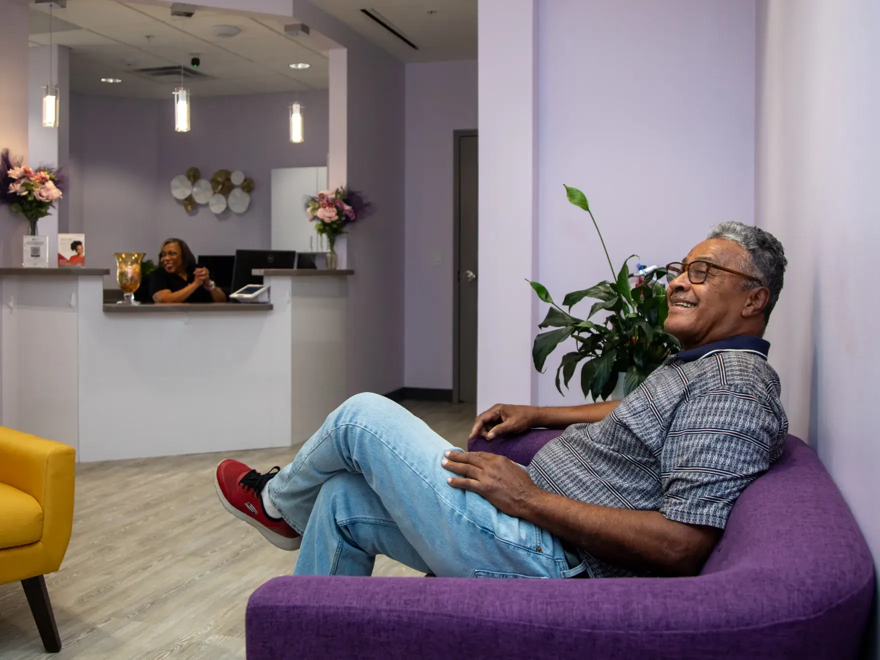 A man sits on a purple couch, smiling, while a receptionist works at a desk in the background.