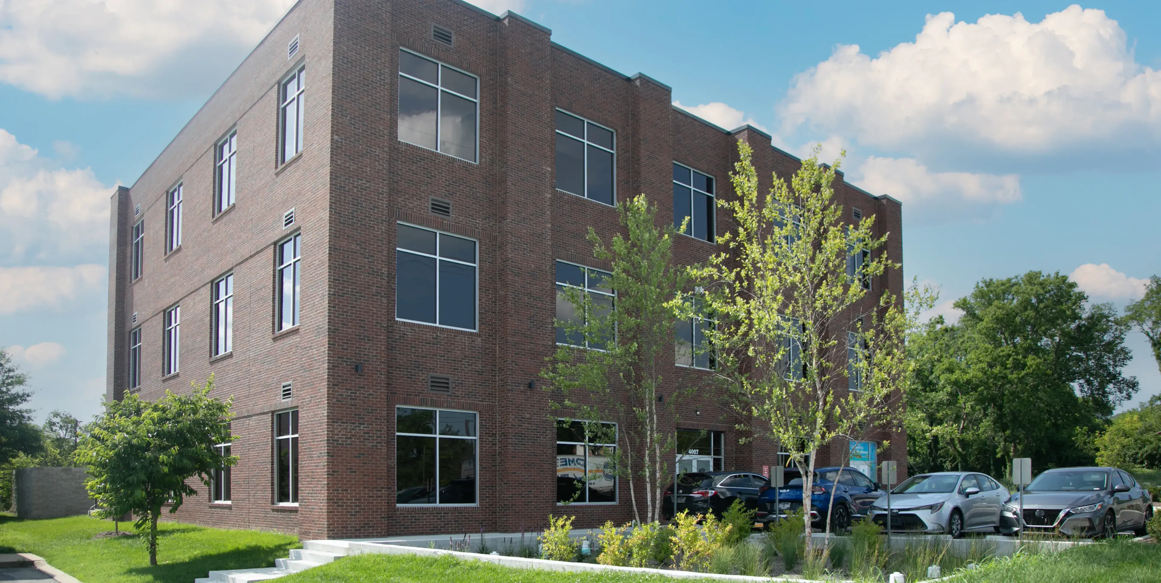 A three-story brick office building with large windows, surrounded by trees and parked cars.
