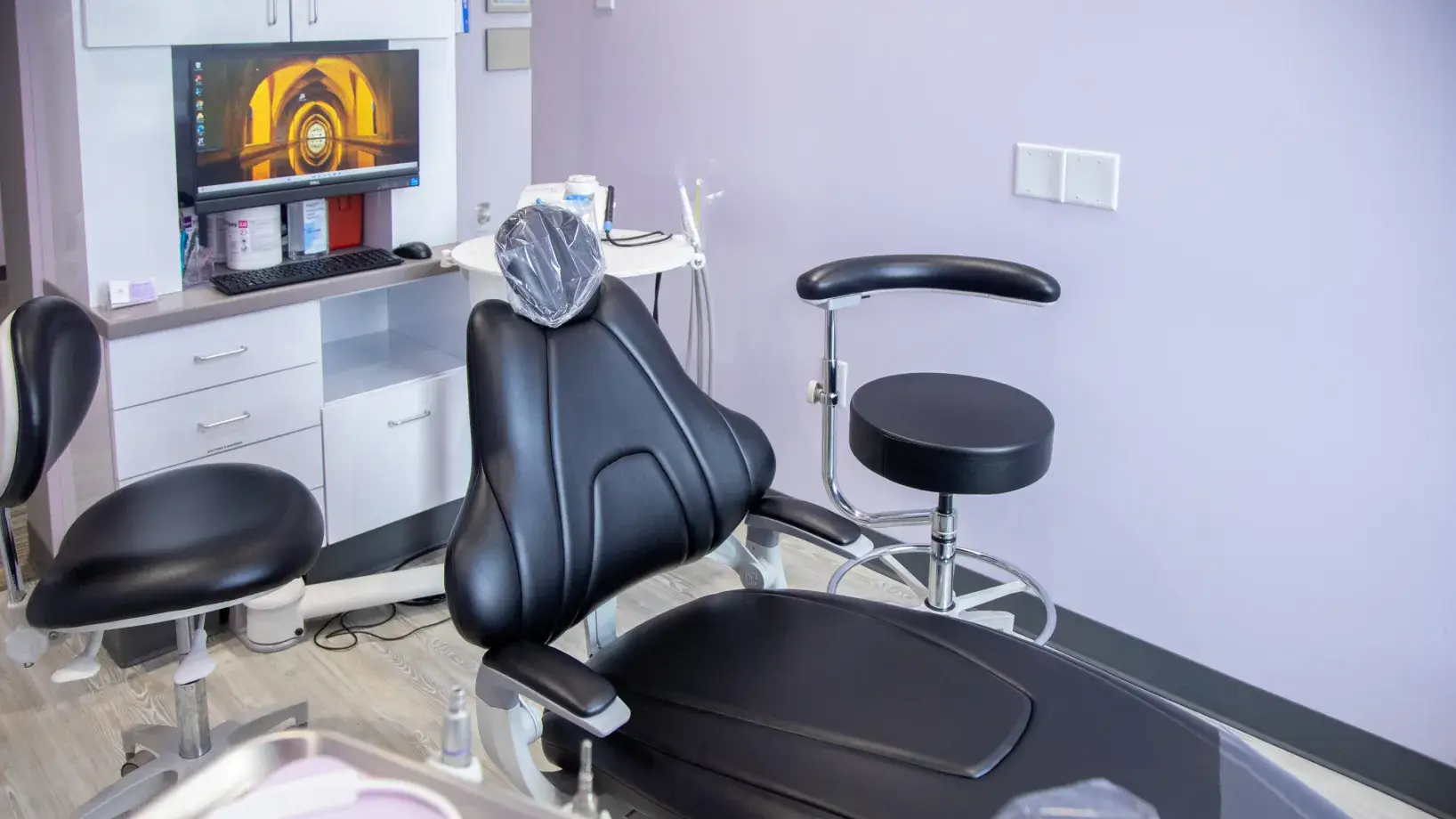 Empty dental examination room with a black dental chair and a computer on a desk.