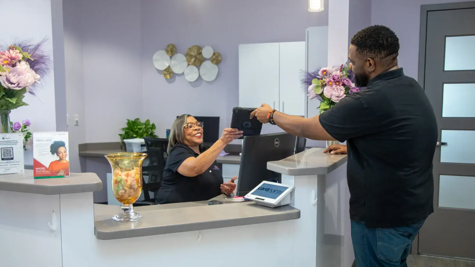 A man hands a tablet to a smiling woman at a reception desk.