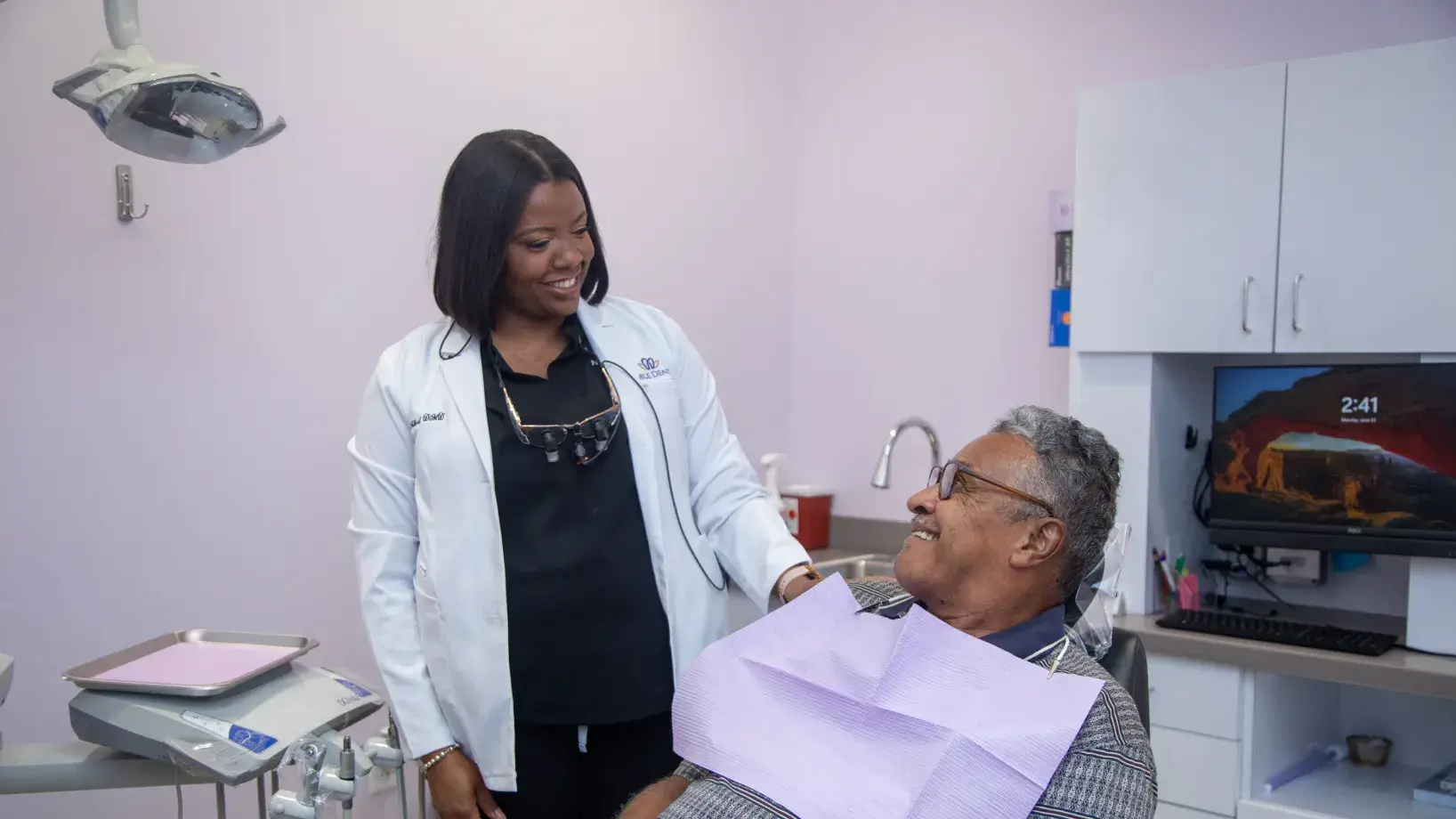 A dentist talks to a smiling patient seated in a dental chair.