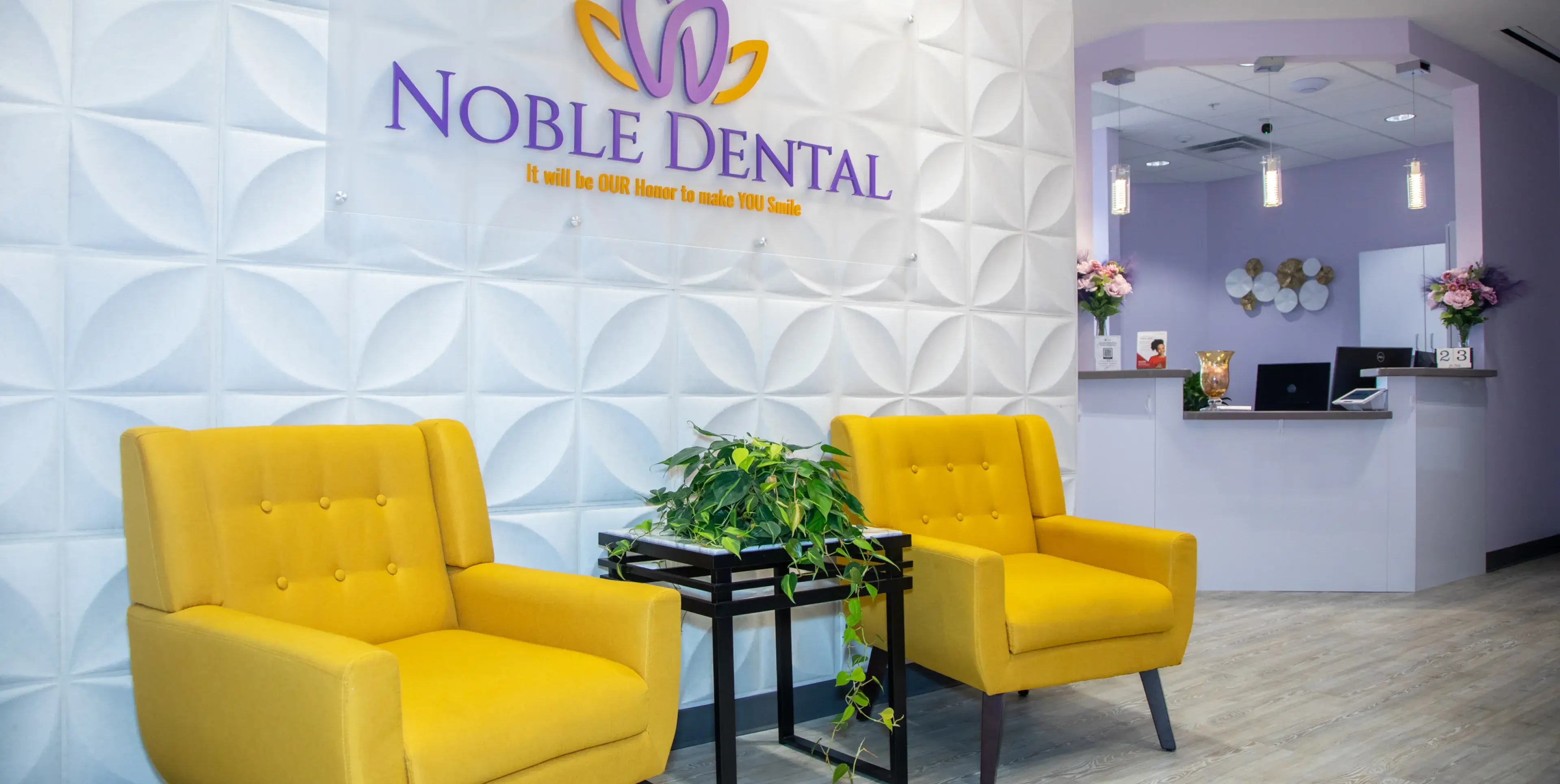 Two yellow chairs and a plant in a dental office waiting area with a reception desk nearby.