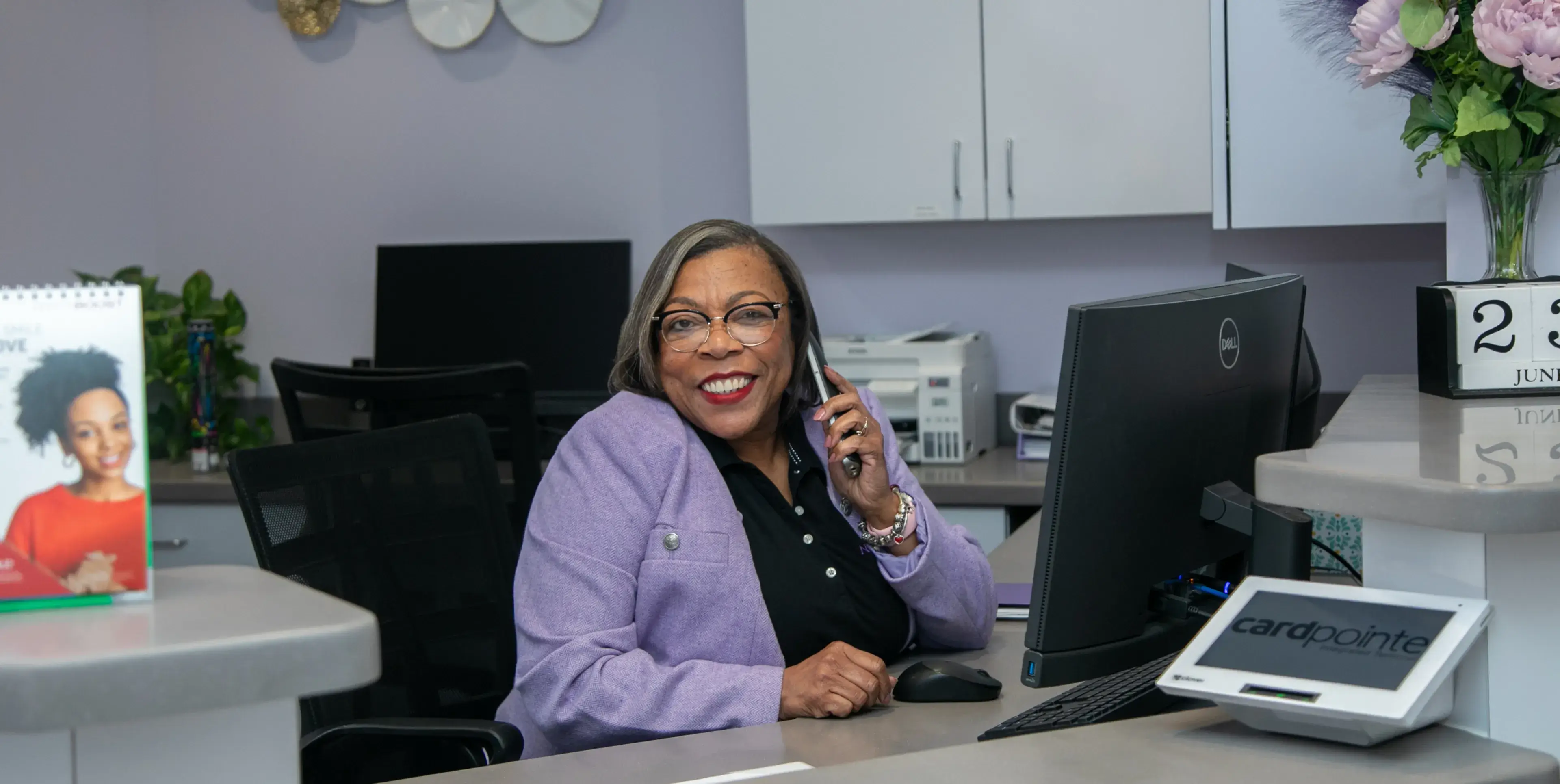 Smiling woman in purple jacket talks on phone at office desk with computer.