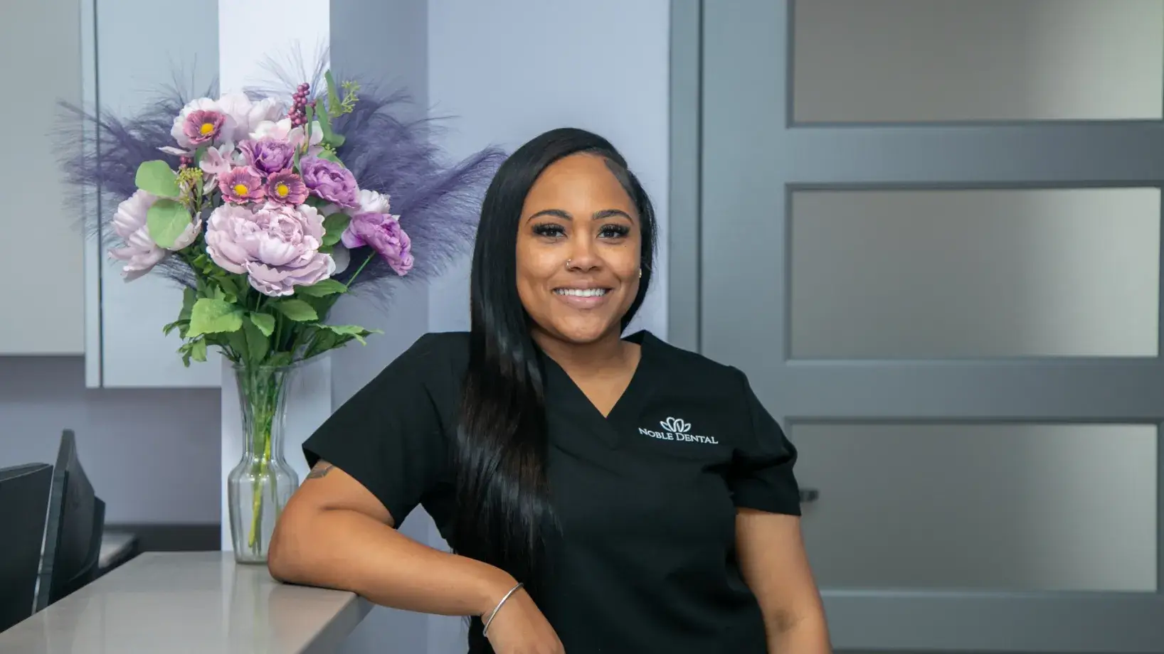 A person in dental scrubs smiling and leaning on a counter next to a vase of flowers.