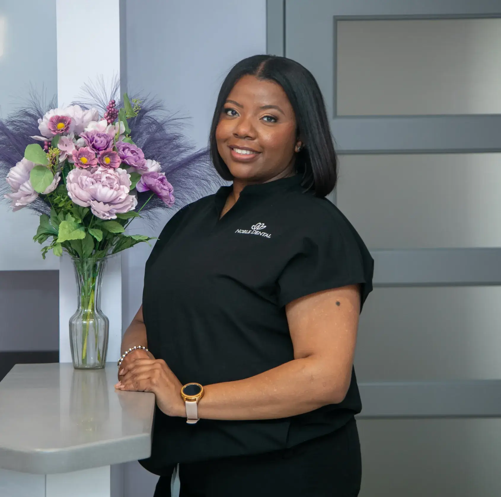 A person in a black shirt smiles while standing next to a vase of purple flowers.