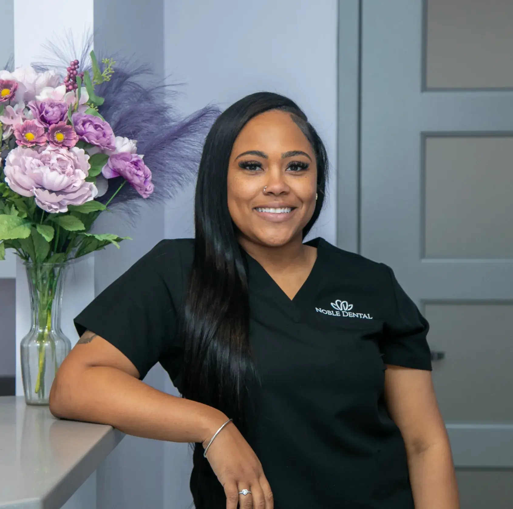 Smiling woman in black scrubs stands by a counter with flowers in the background.