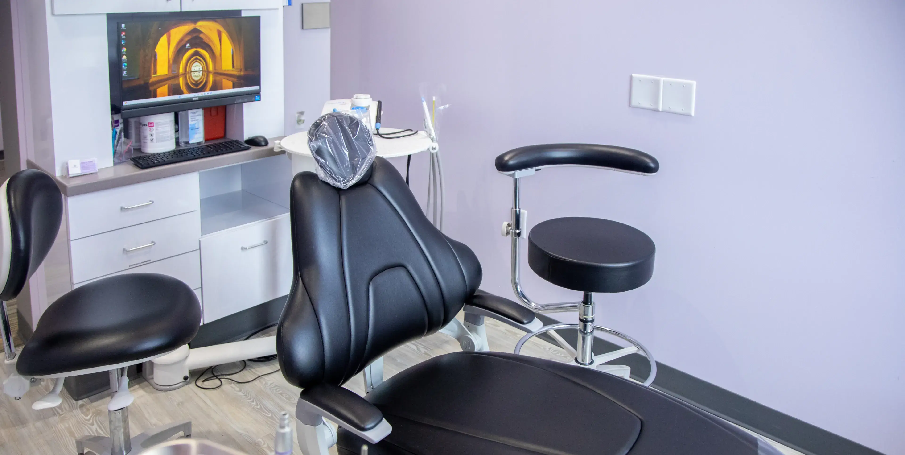 A dental examination room with a black chair, a computer screen, and various dental tools.