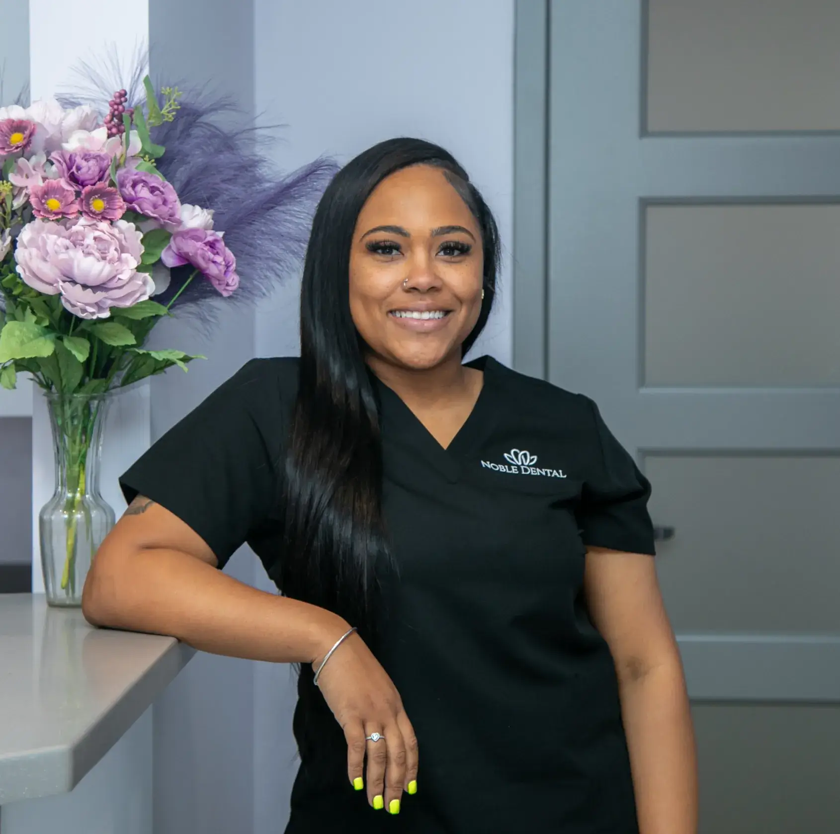 A woman in a black uniform smiles, standing beside a counter with a flower vase.