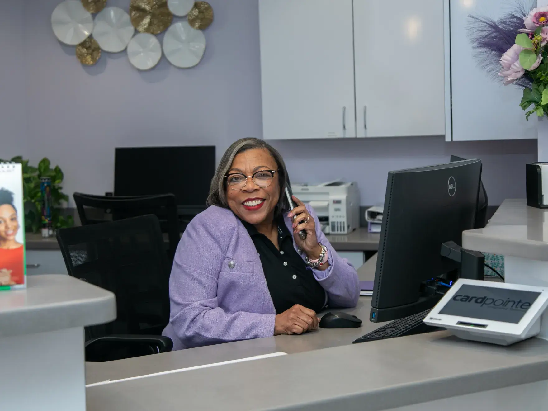A woman is smiling while talking on the phone at an office reception desk.
