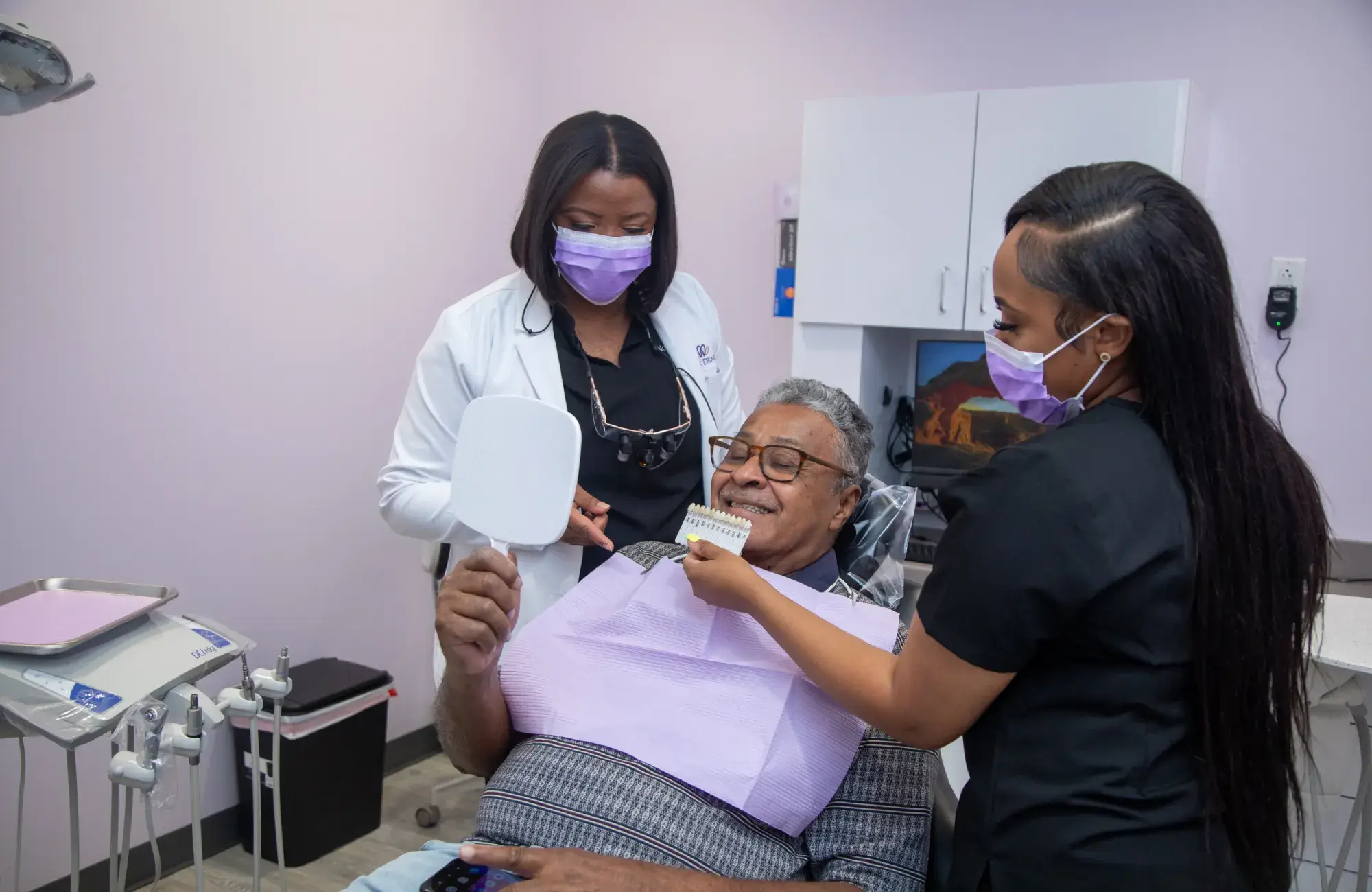 A dentist and assistant help a man check his teeth using a mirror and shade guide.