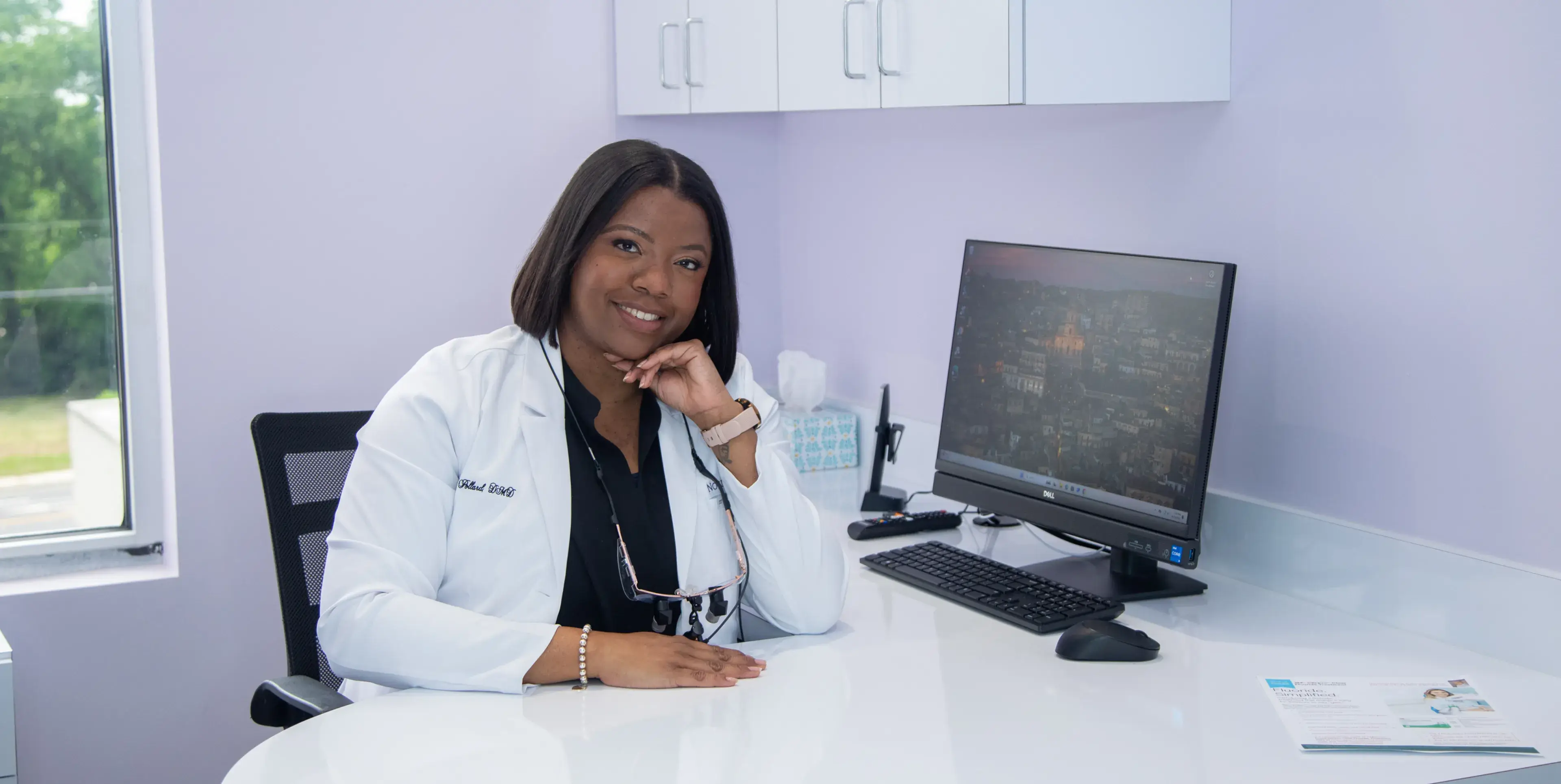 A woman in a white coat smiles, sitting at a desk with a computer in an office.