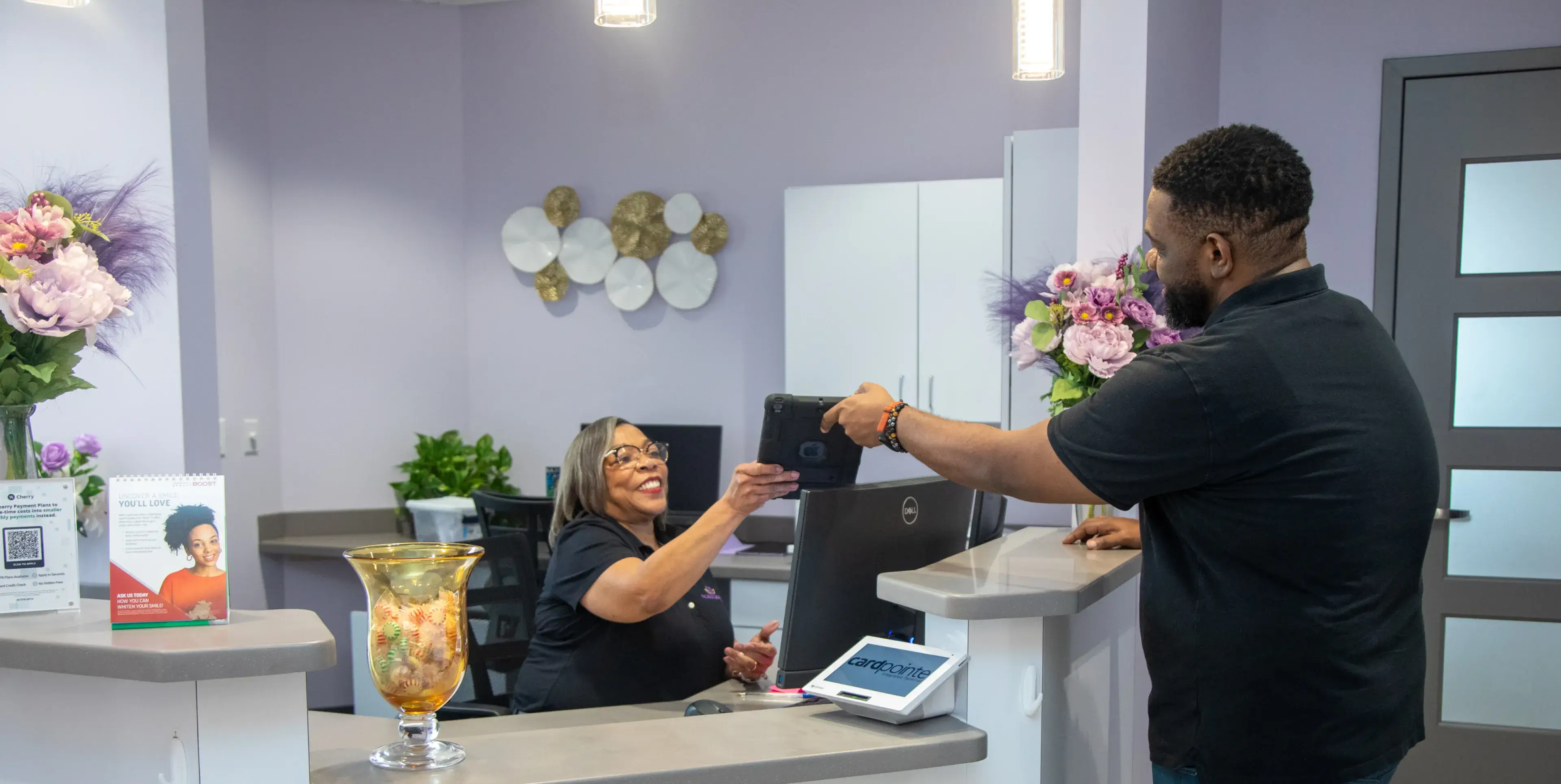 A man hands a tablet to a smiling receptionist at a desk in an office.