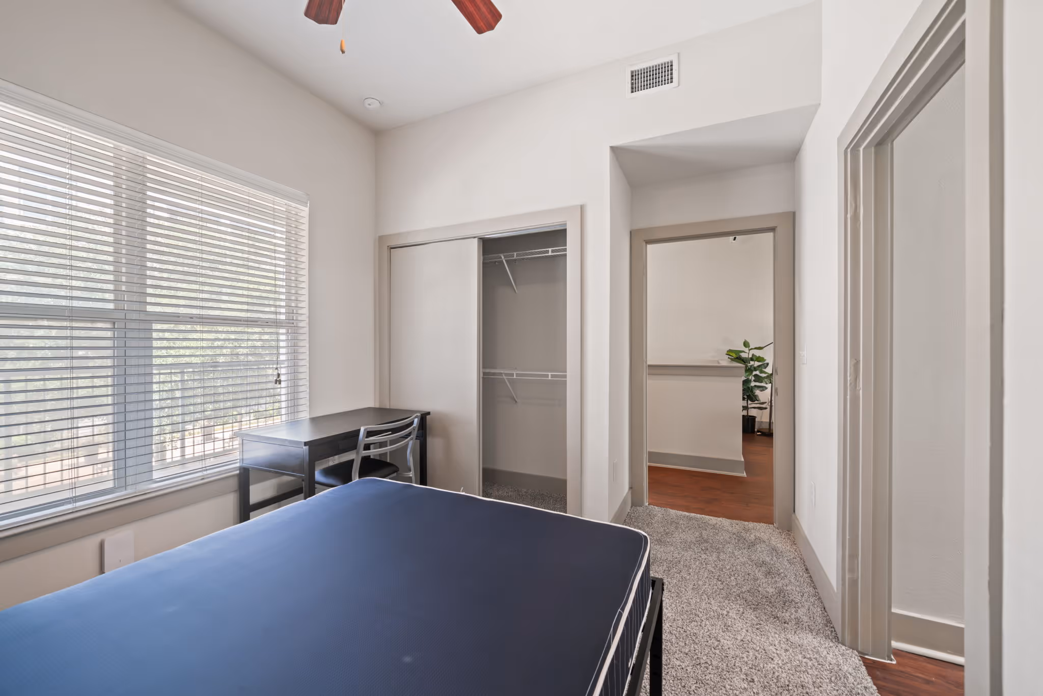 View of an apartment bedroom showing a private workspace, mattress, large windows, and access to the hallway.