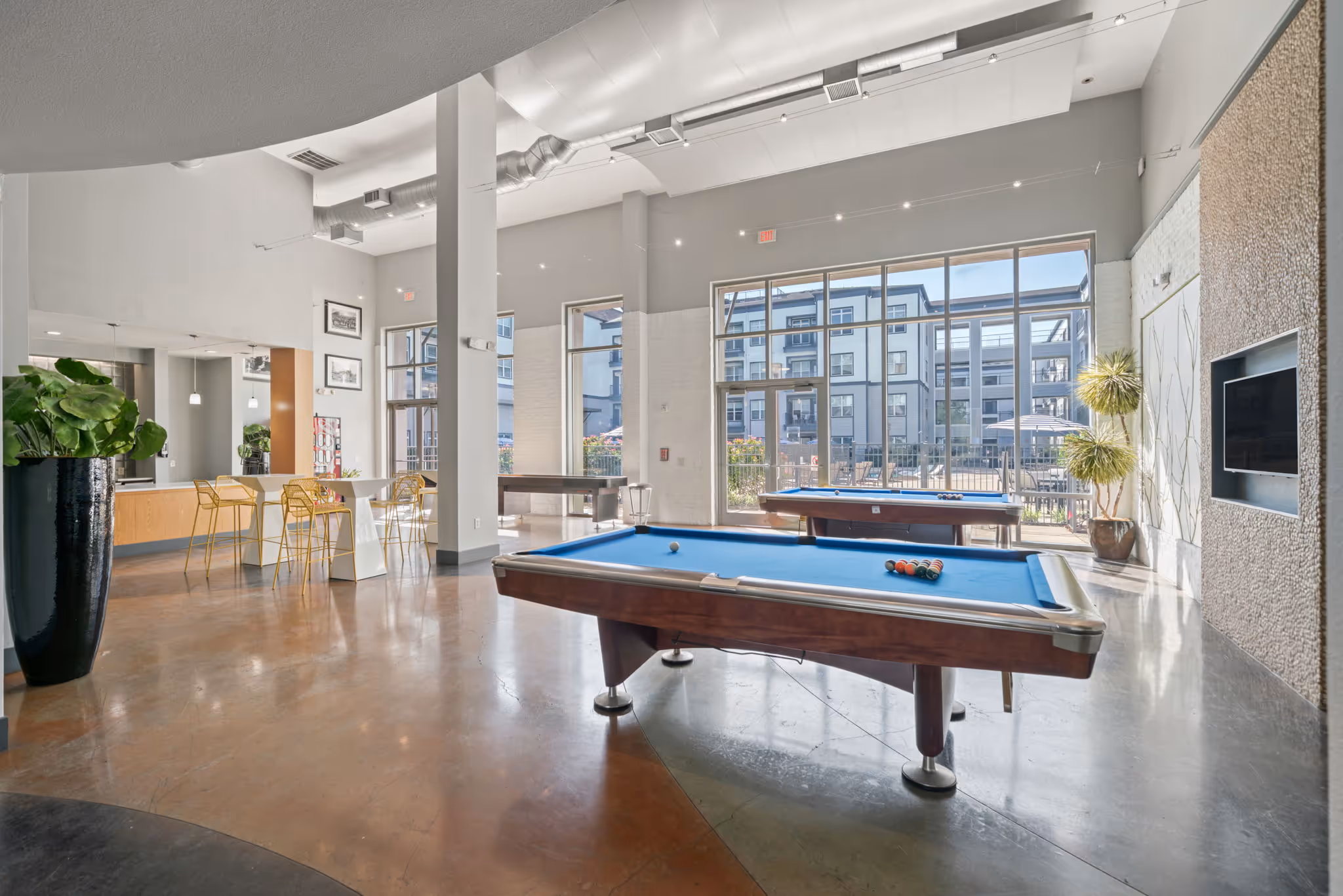 Spacious community lounge featuring two blue-felt billiard tables, high-top seating, large windows overlooking the pool, and modern polished concrete flooring at The Lofts at Wolf Pen Creek in College Station, TX