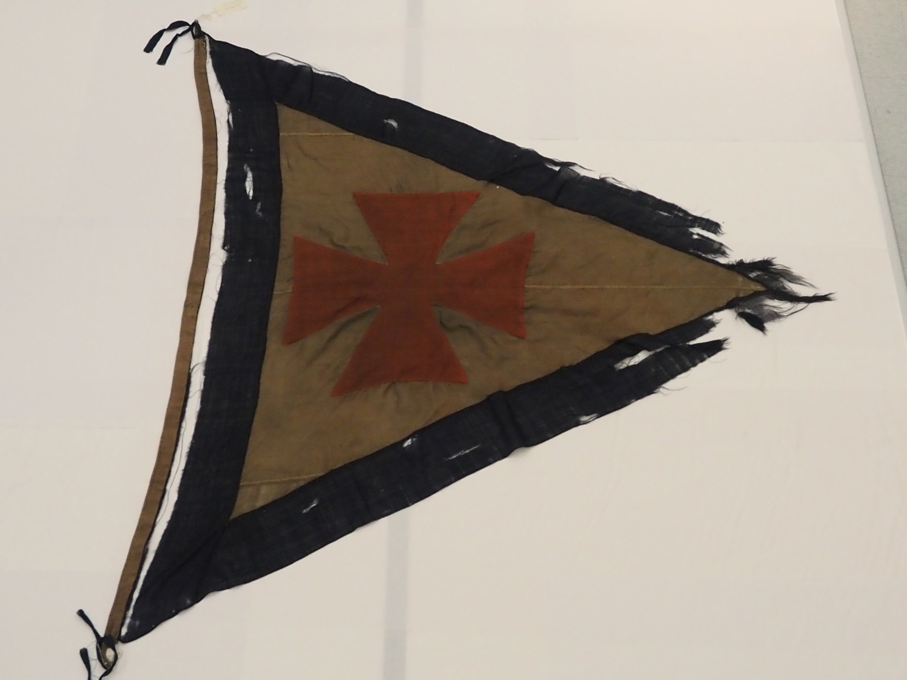 Frayed triangular flag with a red cross centered on a faded tan background, edged with a dark border, showing signs of age and wear.