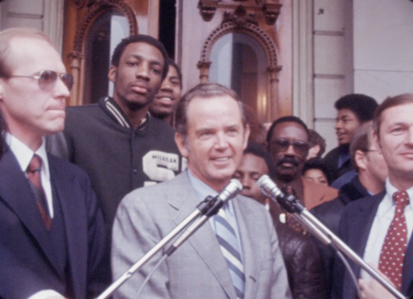 A man in a suit speaks at microphones outside a building, surrounded by a crowd of men standing behind him.