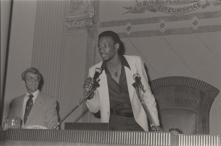 A man stands at a podium holding a microphone and speaking, while another man stands nearby, in an ornate indoor chamber.