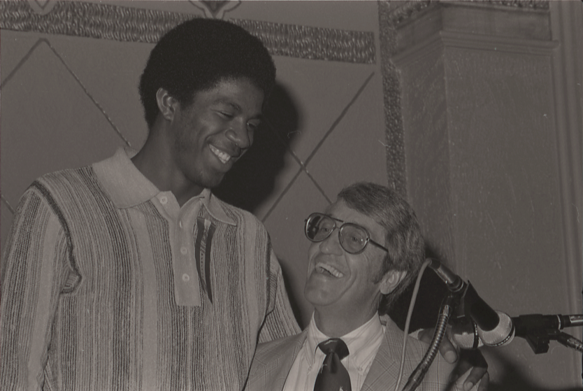 A tall man smiles down at a man with glasses near a microphone, both appearing to share a light moment in an indoor setting.