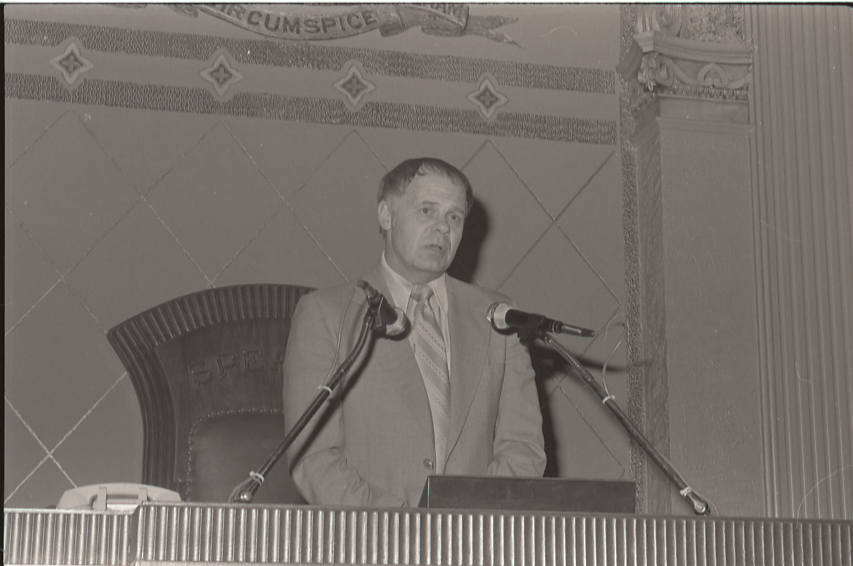 A man in a suit speaks at a podium with two microphones in an ornate indoor chamber.
